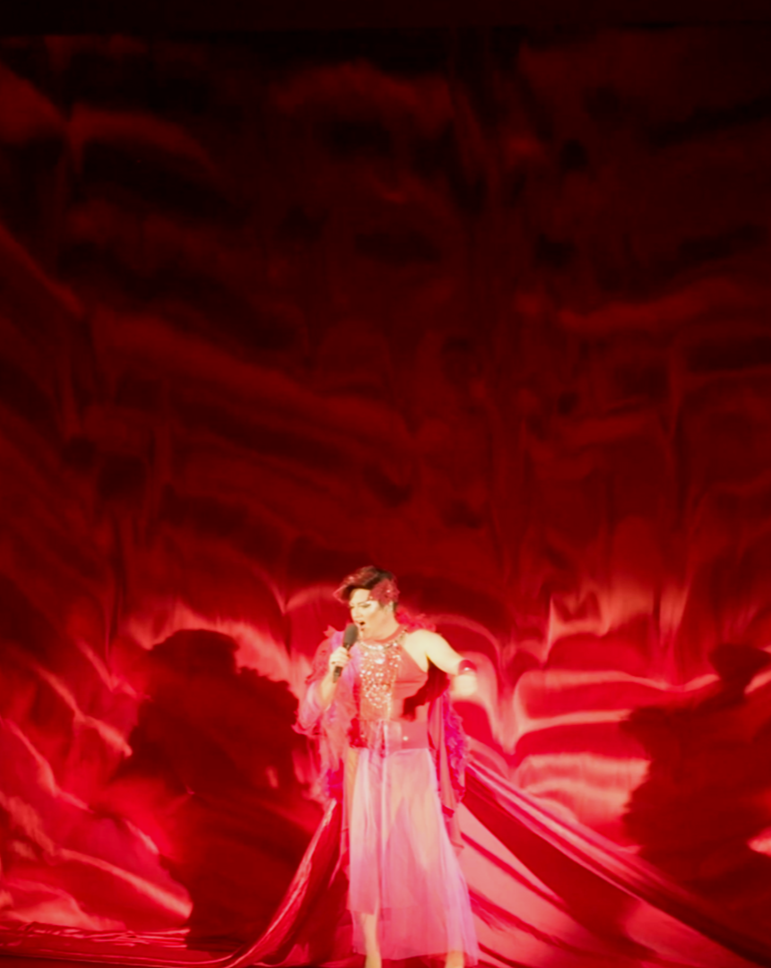 Philip Richart, verkleidet als Lulu Mimeuse, in einem pinken Kleid auf einer Bühne vor rotem Hintergrund, hält Mikrofon in der Hand und singt.