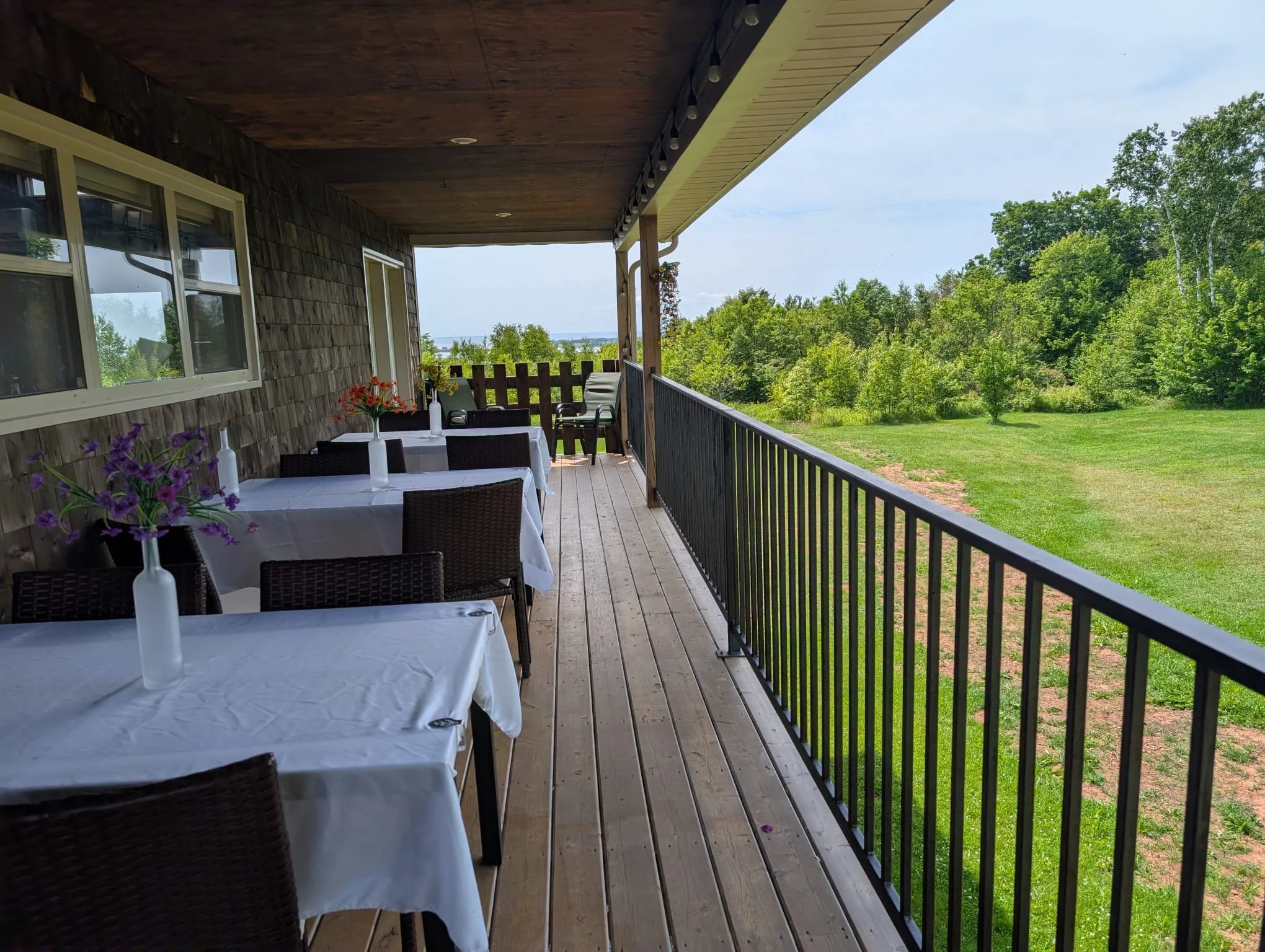 Image of a wooden porch or balcony with a table set for outdoor dining, decorated with flower vases. Beyond the porch, a lush green grassy yard and trees are visible under a partly cloudy sky.