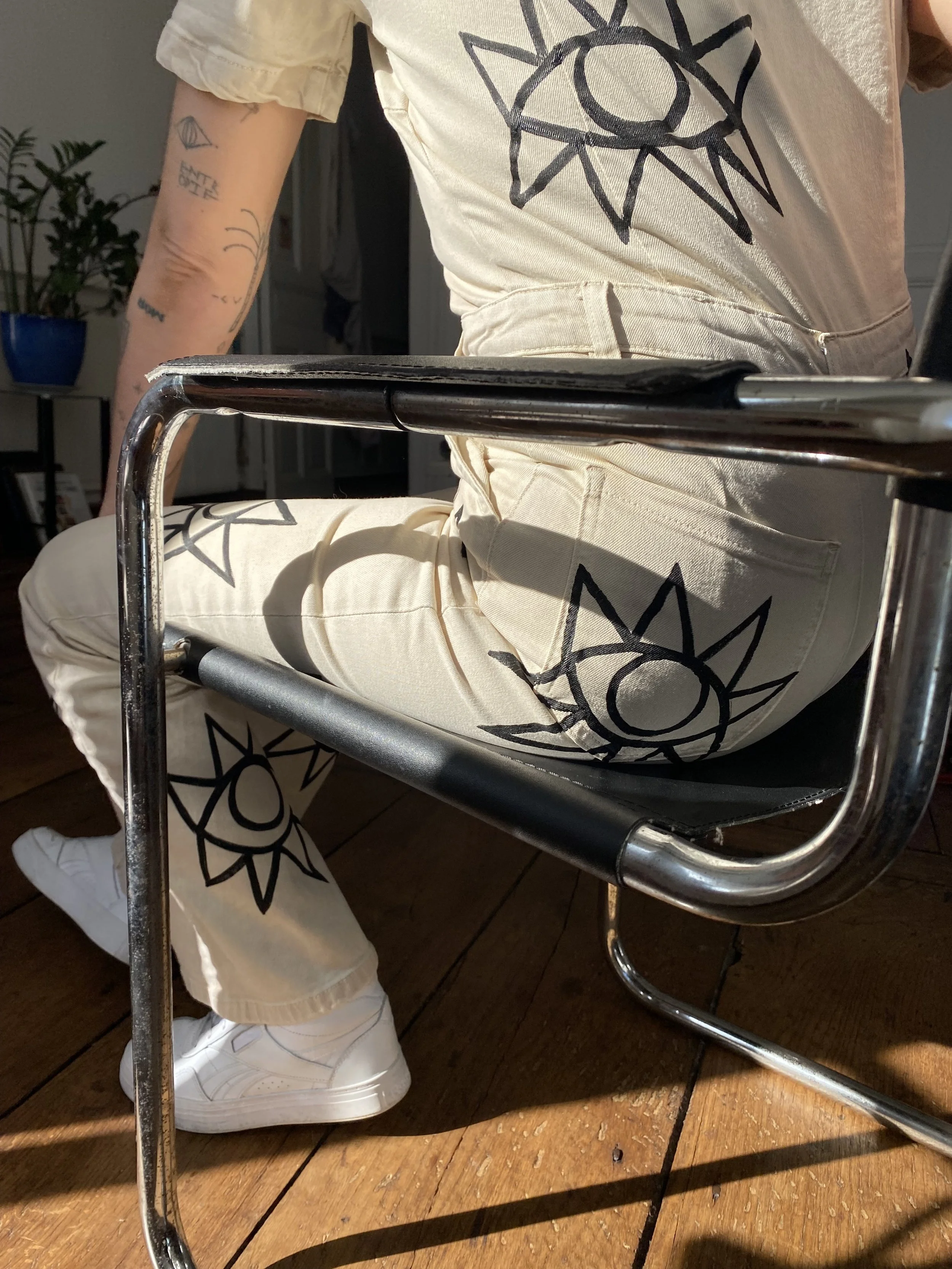 Person sitting on a chair with a glass top, wearing cream-colored pants and a T-shirt with a sun and eye design, white sneakers, and has visible tattoos on their arm. The setting is indoors with wooden flooring and a potted plant in the background.