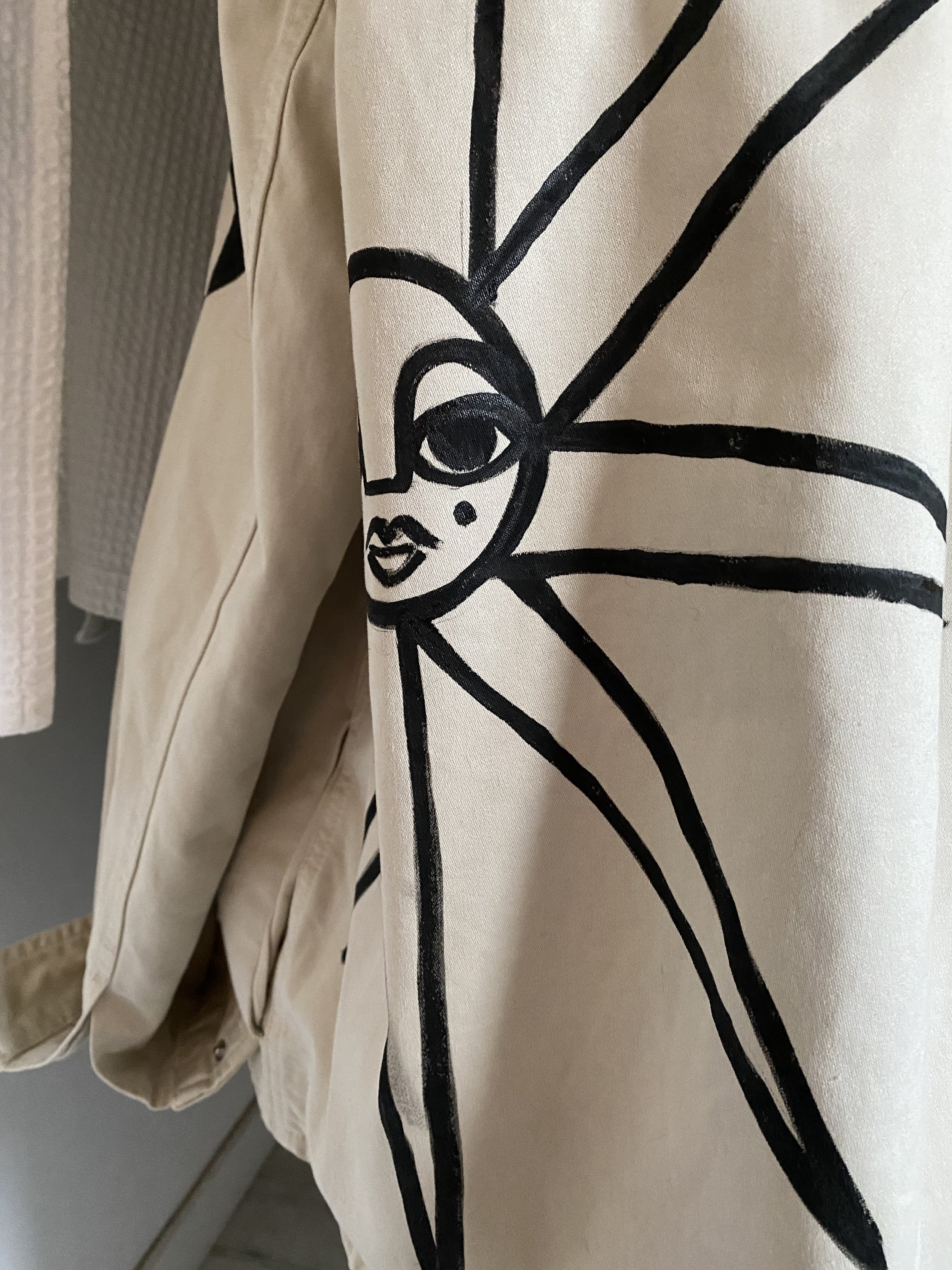 A beige or cream-colored jacket with a black line art design of a face with one eye, lips, and a small dot, with radiating lines around the face.