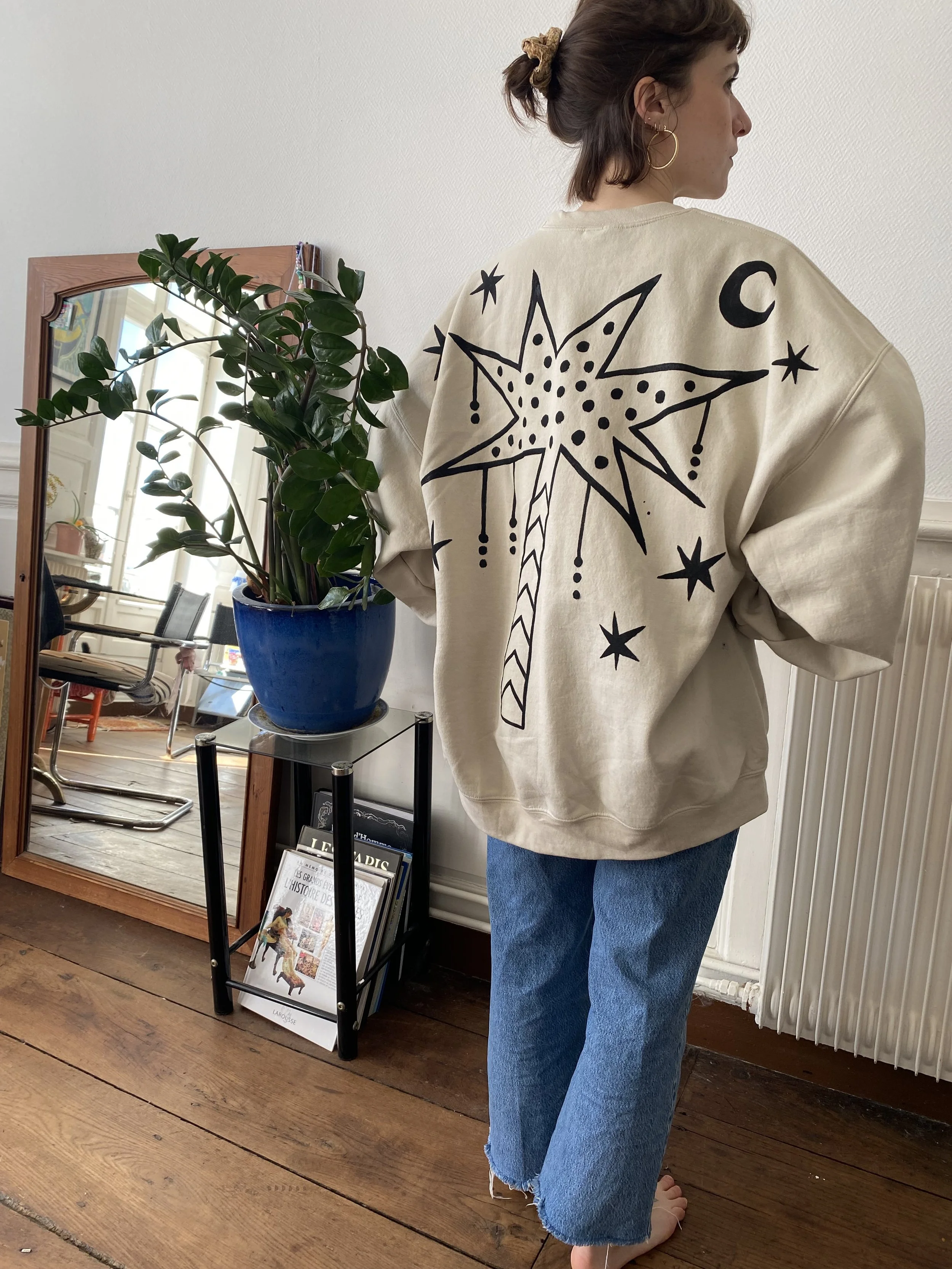 A woman standing with her back to the camera, wearing a cream sweatshirt with a large black star and celestial designs on the back, and blue jeans. She has a bun hairstyle secured with a scrunchie and is wearing hoop earrings. She is indoors near a l