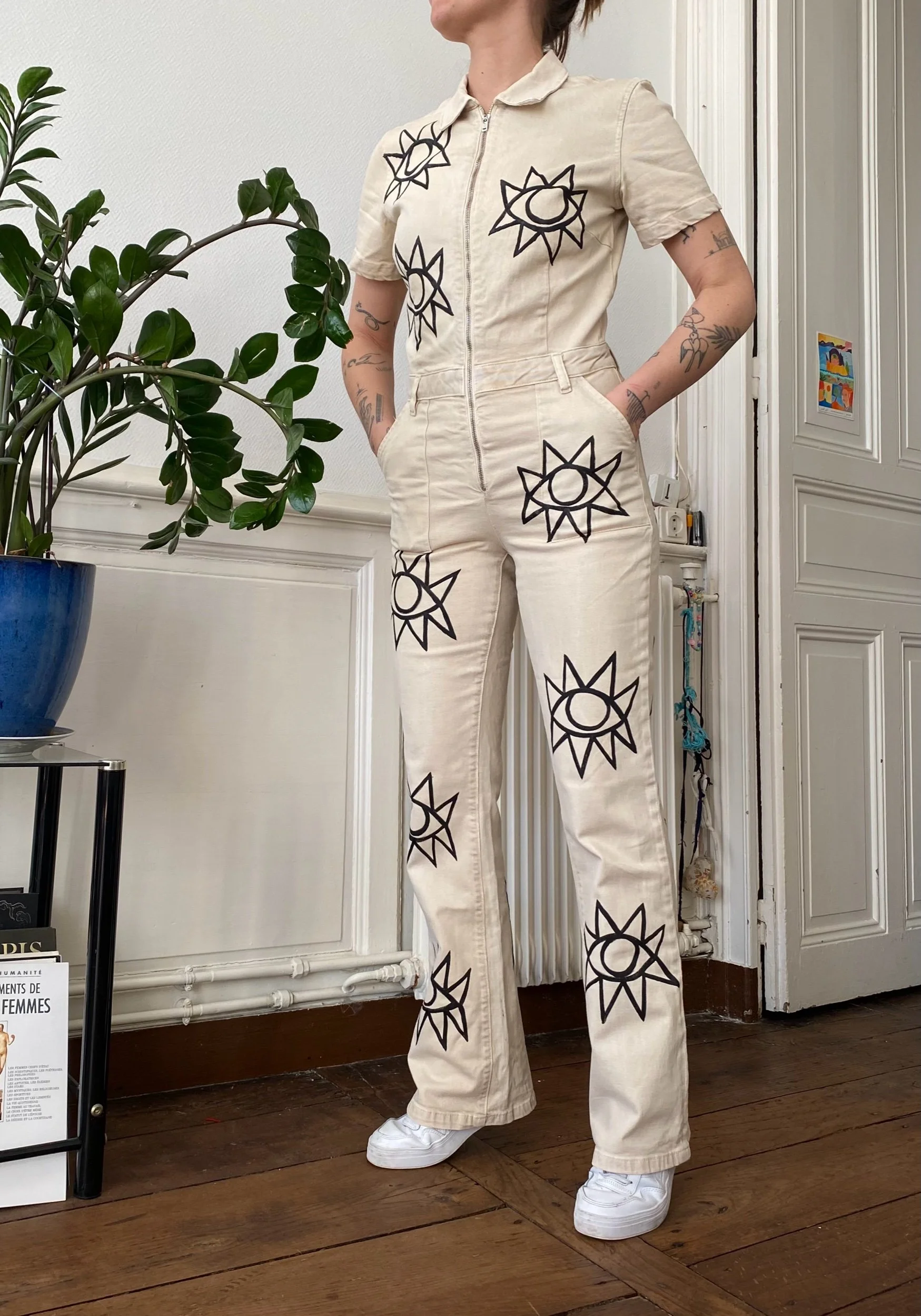 Person wearing a beige jumpsuit with black line art sun and moon designs in a room with wooden floor, white paneled walls, and a blue potted plant.