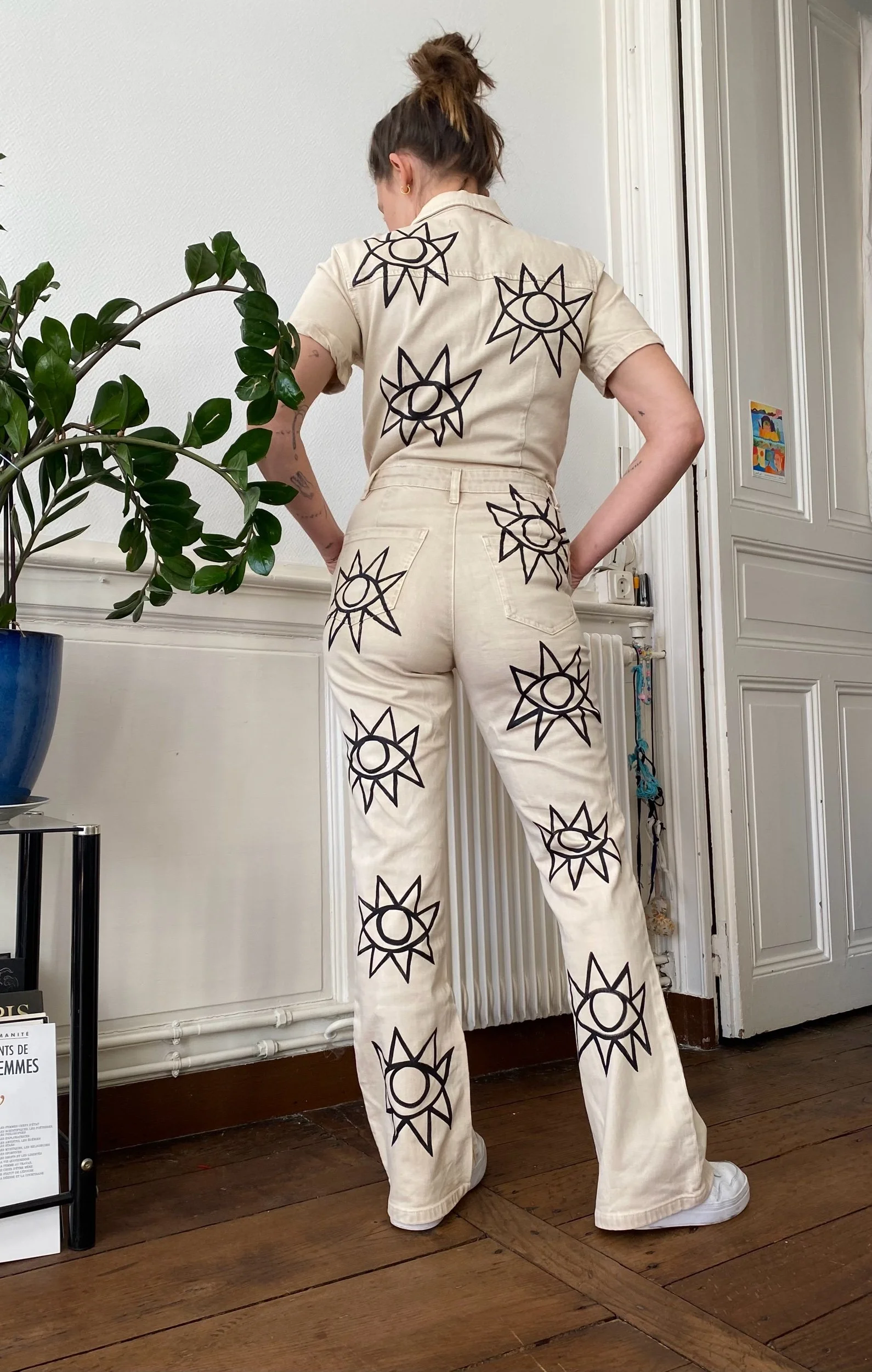 Woman standing indoors, back to the camera, wearing a matching beige shirt and pants with black geometric eye and sun designs, next to a potted plant and a white door.