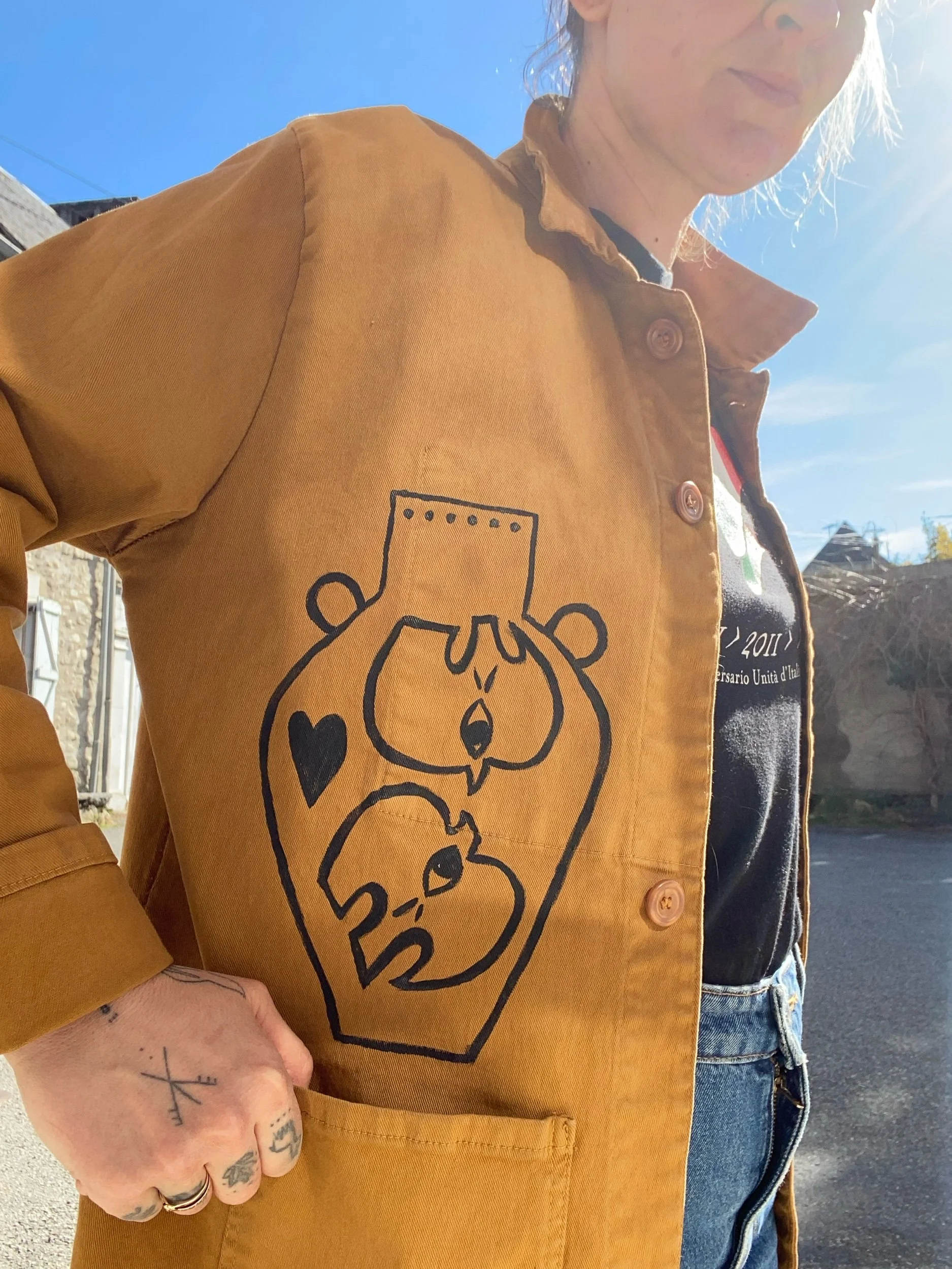 Person wearing a brown jacket with a black and white cartoon bear design on the front, standing outside on a sunny day.