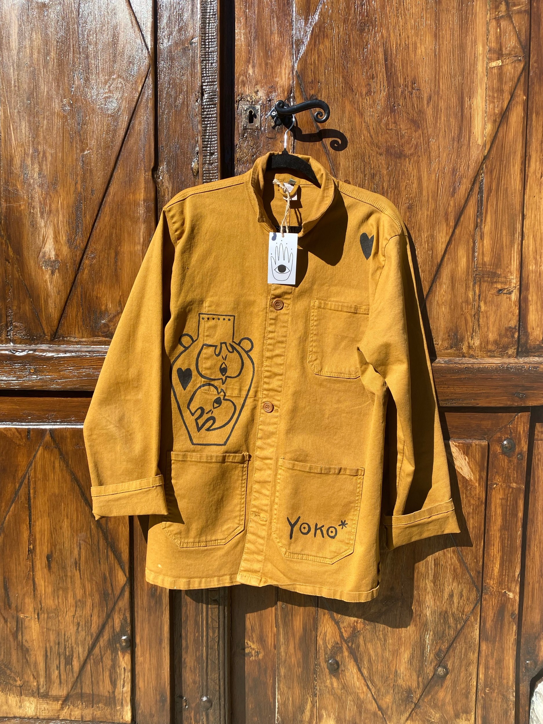Mustard yellow long-sleeve coat hanging on a hook on wooden doors, with hand-drawn black outlines of a bear in a vase and small heart shapes, and the name "Yoko" written on the lower pocket.