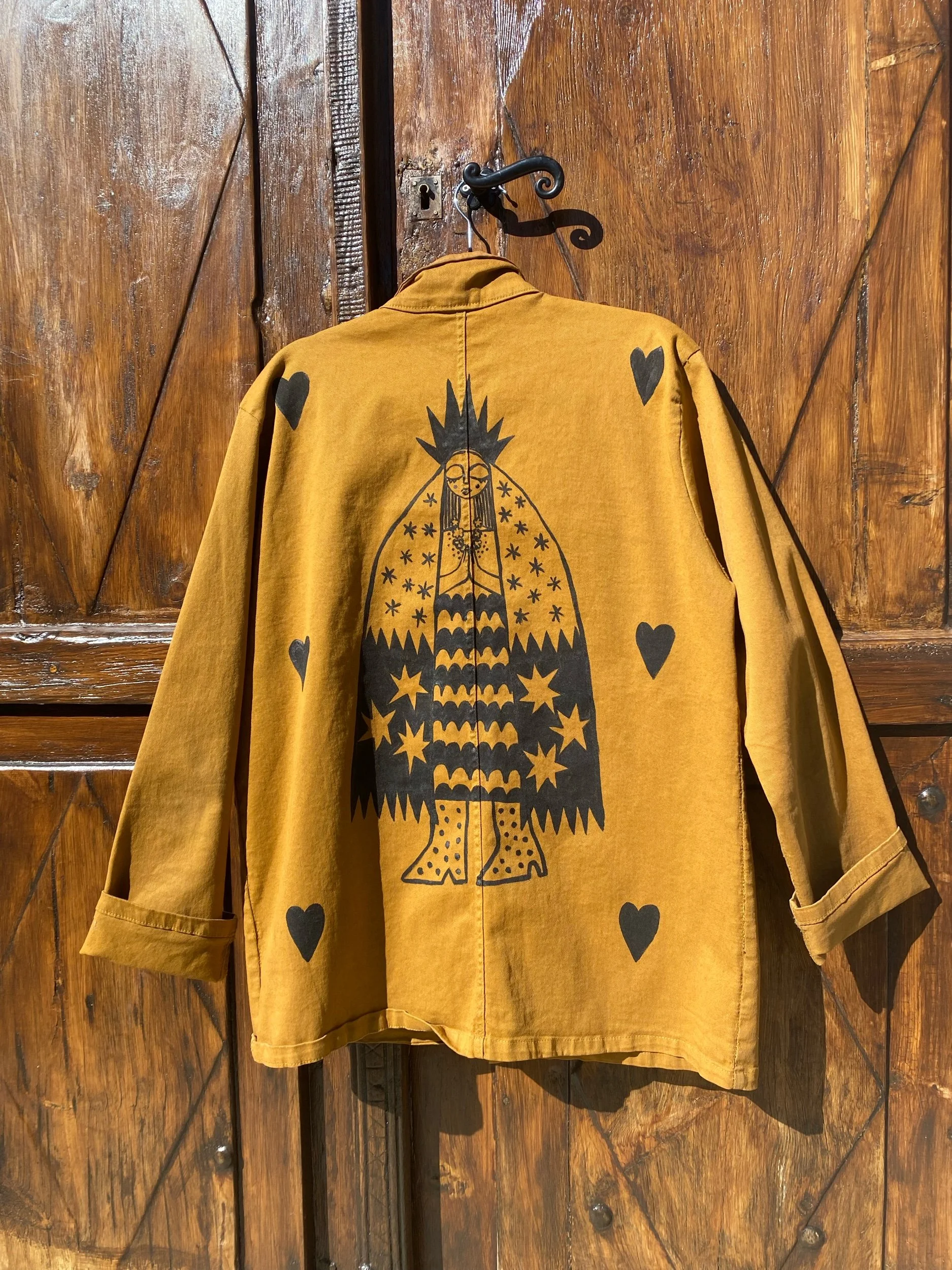 A mustard yellow jacket with a large black graphic of a mystical figure with a headdress and outstretched hands, surrounded by black hearts and stars, hanging on a wooden door.