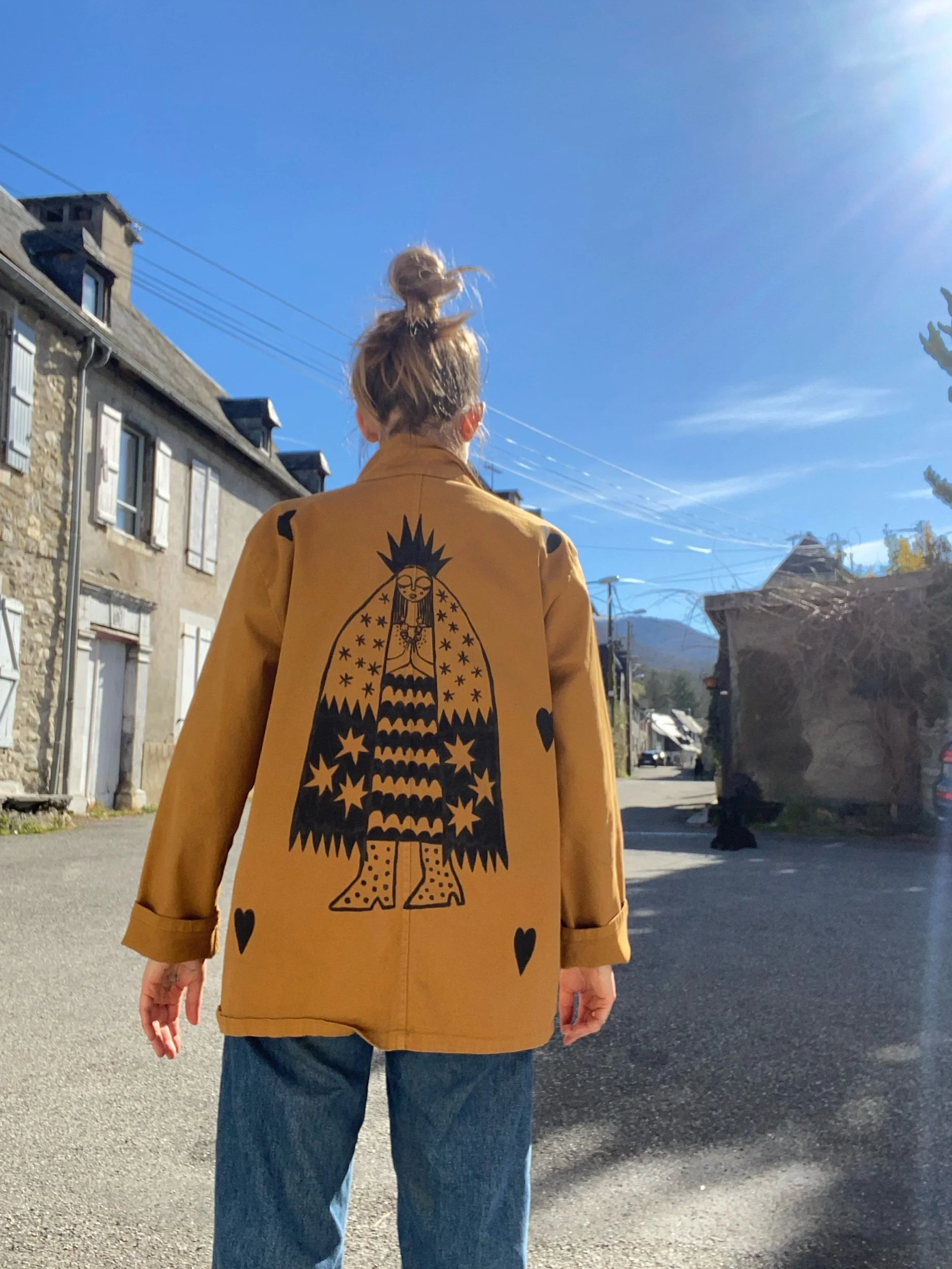 Back view of a person with blonde hair in a top bun, wearing a mustard yellow jacket with a large black graphic of a crowned woman and hearts on the back, and blue jeans, standing on a street under a clear blue sky.