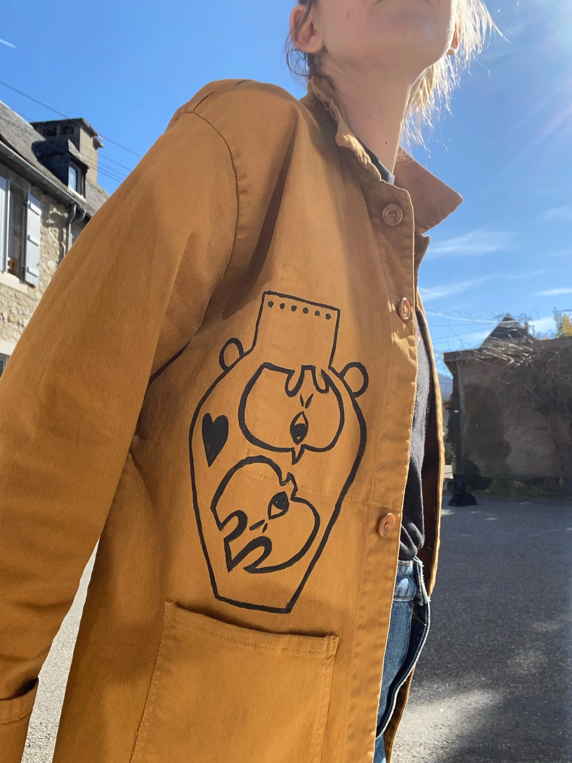 Person wearing a tan jacket with a black line drawing of two cartoon dog faces and a heart on the sleeve. The person is outdoors under a blue sky.
