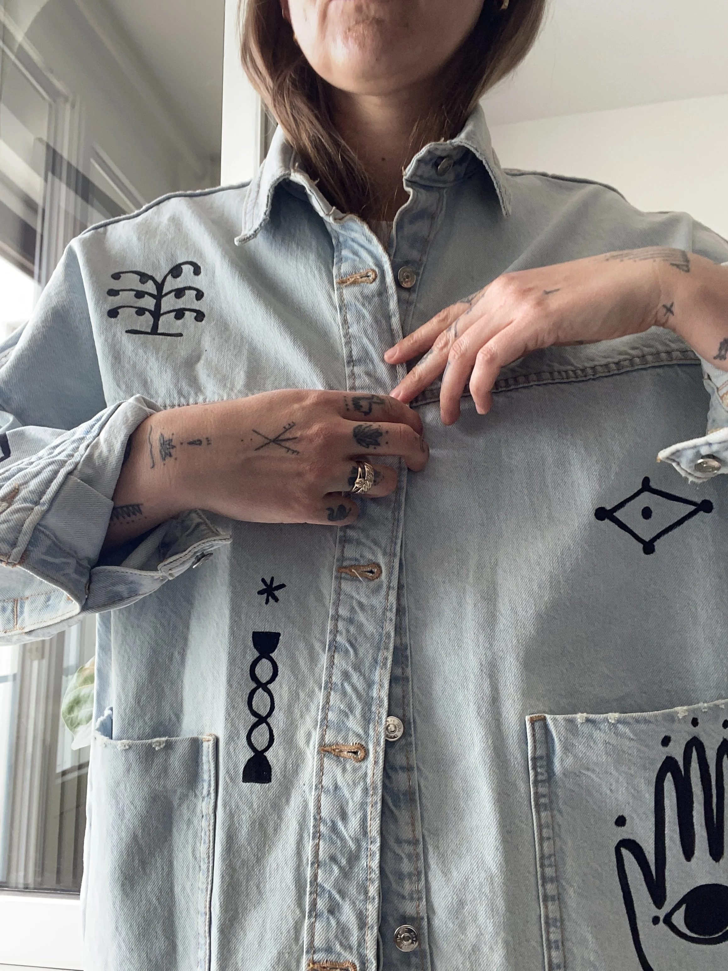 Close-up of a person wearing a light denim jacket with black decorative symbols and patches. The person is holding the collar of the jacket with both hands, revealing tattoos on their fingers and hands. The background shows a window with blinds.