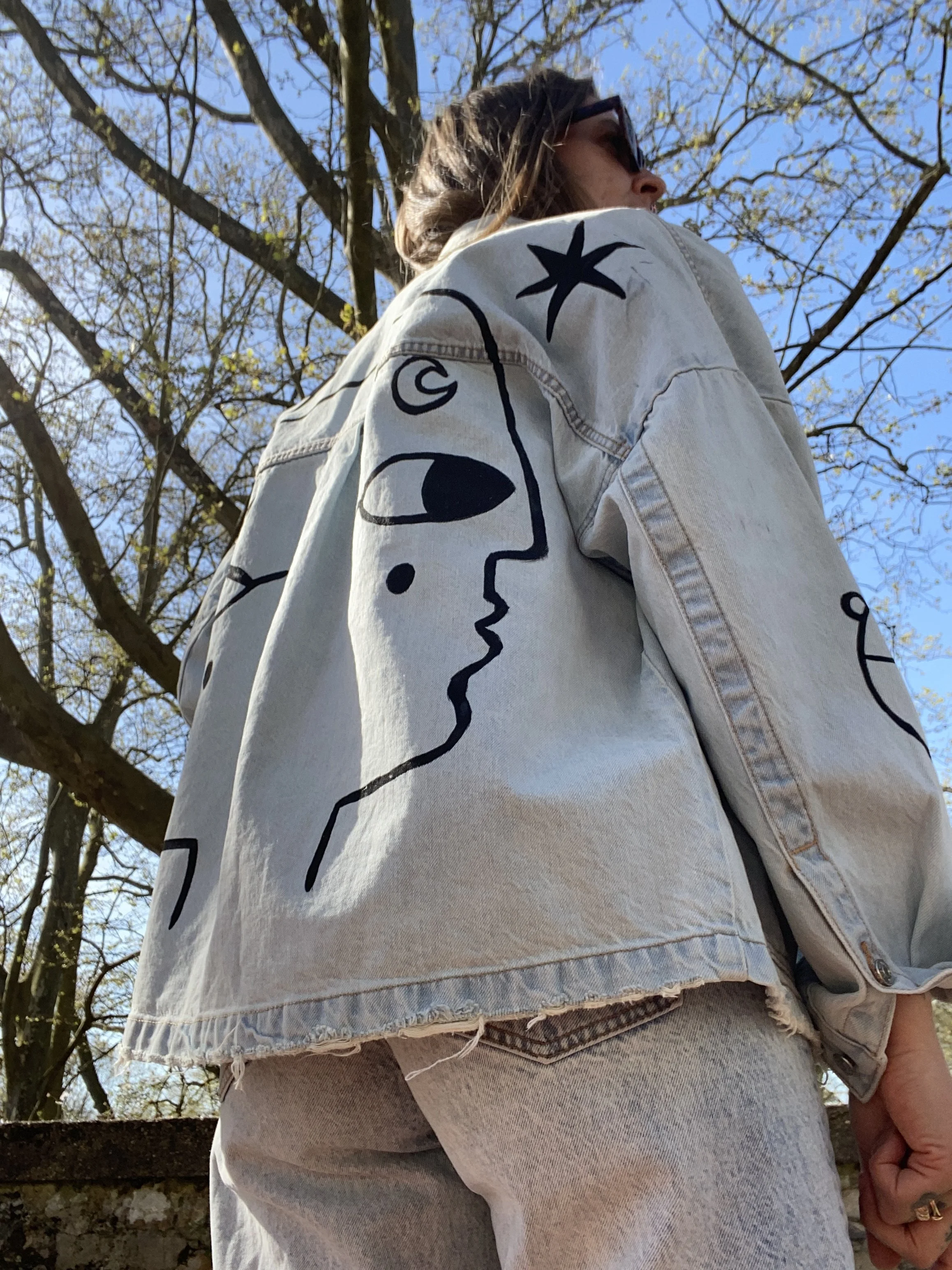 A woman wearing sunglasses and a light denim jacket with abstract face and star graphics, standing outdoors near a tree with budding leaves on a clear sunny day.