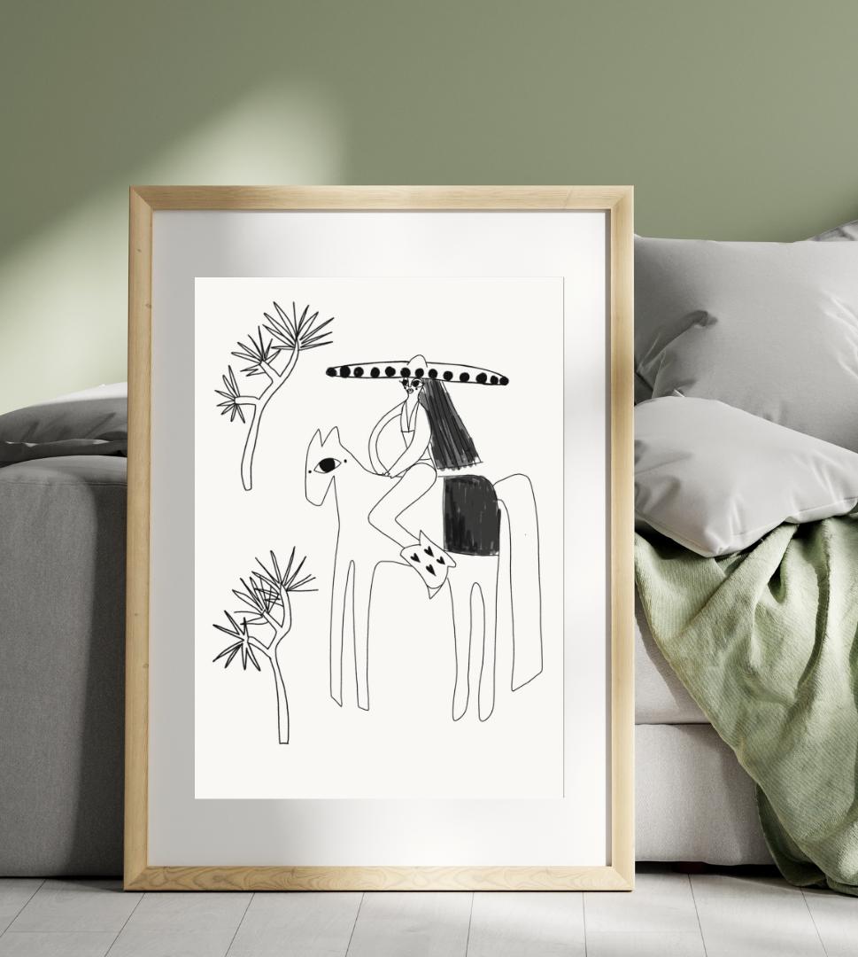 A framed black and white line art drawing of a person riding a horse, wearing a large sombrero, with simple stylized trees in the background, leaning against a bed with gray pillows and a green blanket.