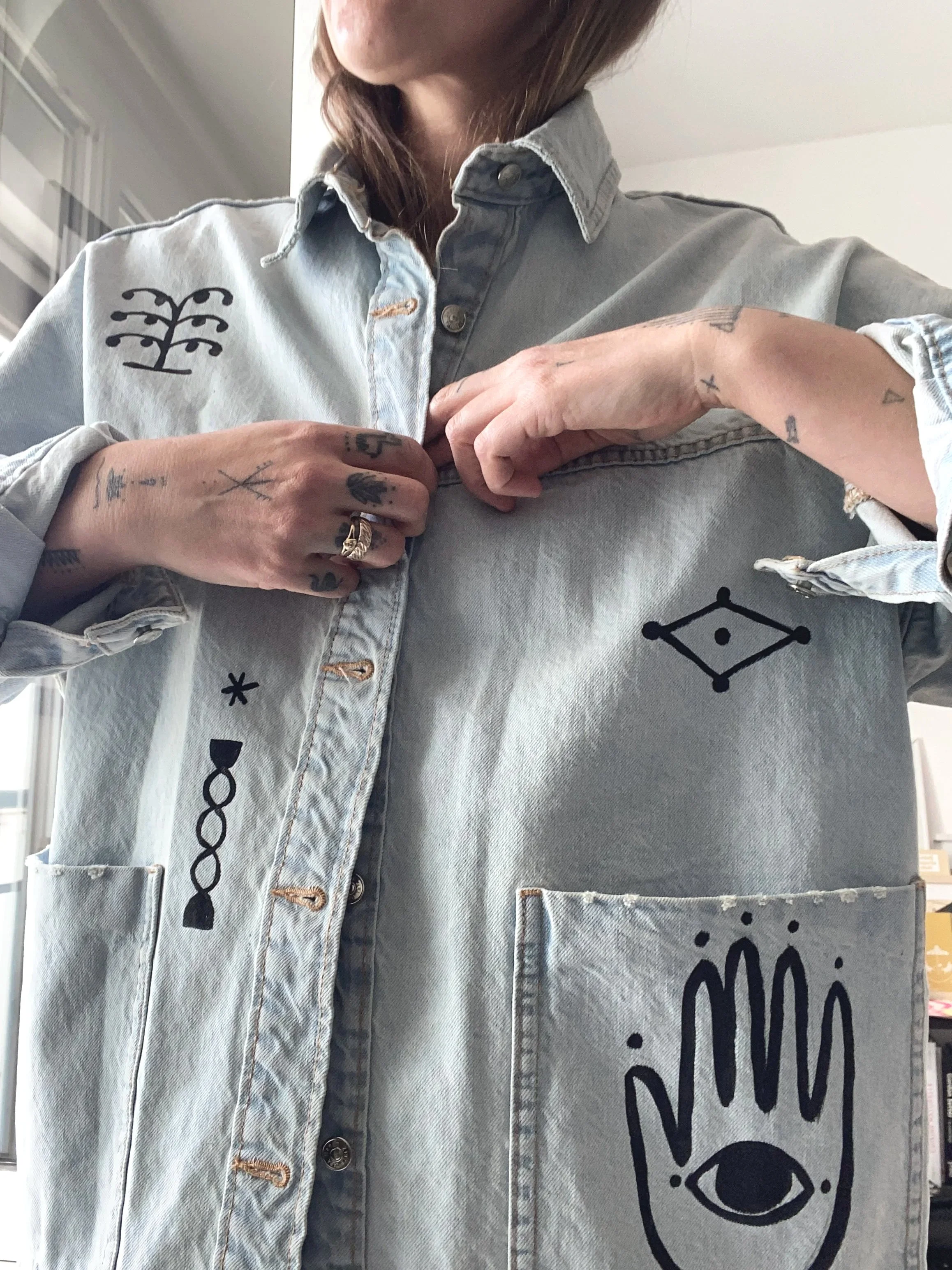 Person wearing a light denim jacket with black hand-drawn symbols and designs, adjusting the buttons on the front.