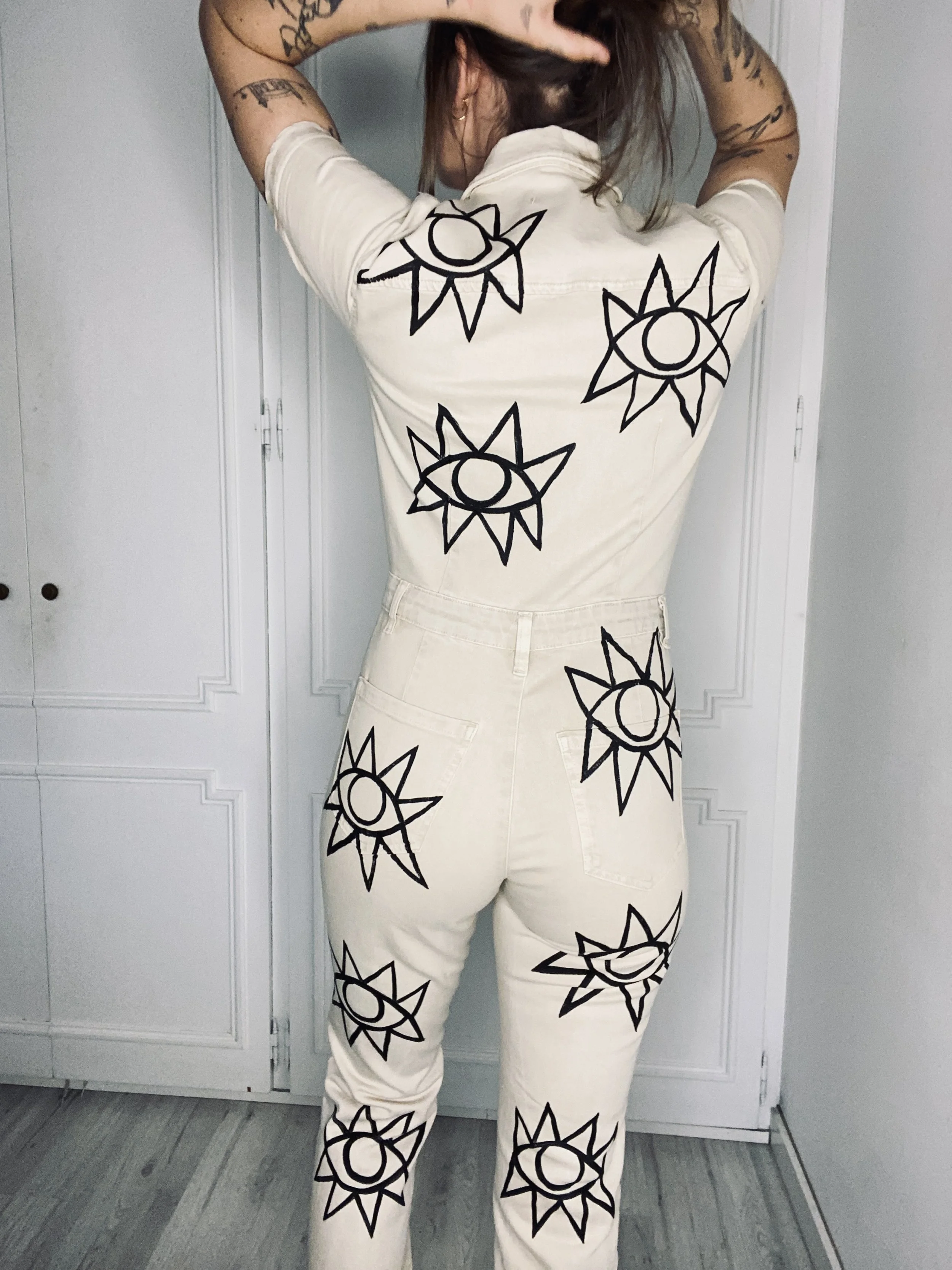 A person with tattoos on their arms is standing with their back to the camera, wearing a beige outfit with black sun and eye designs printed on it.
