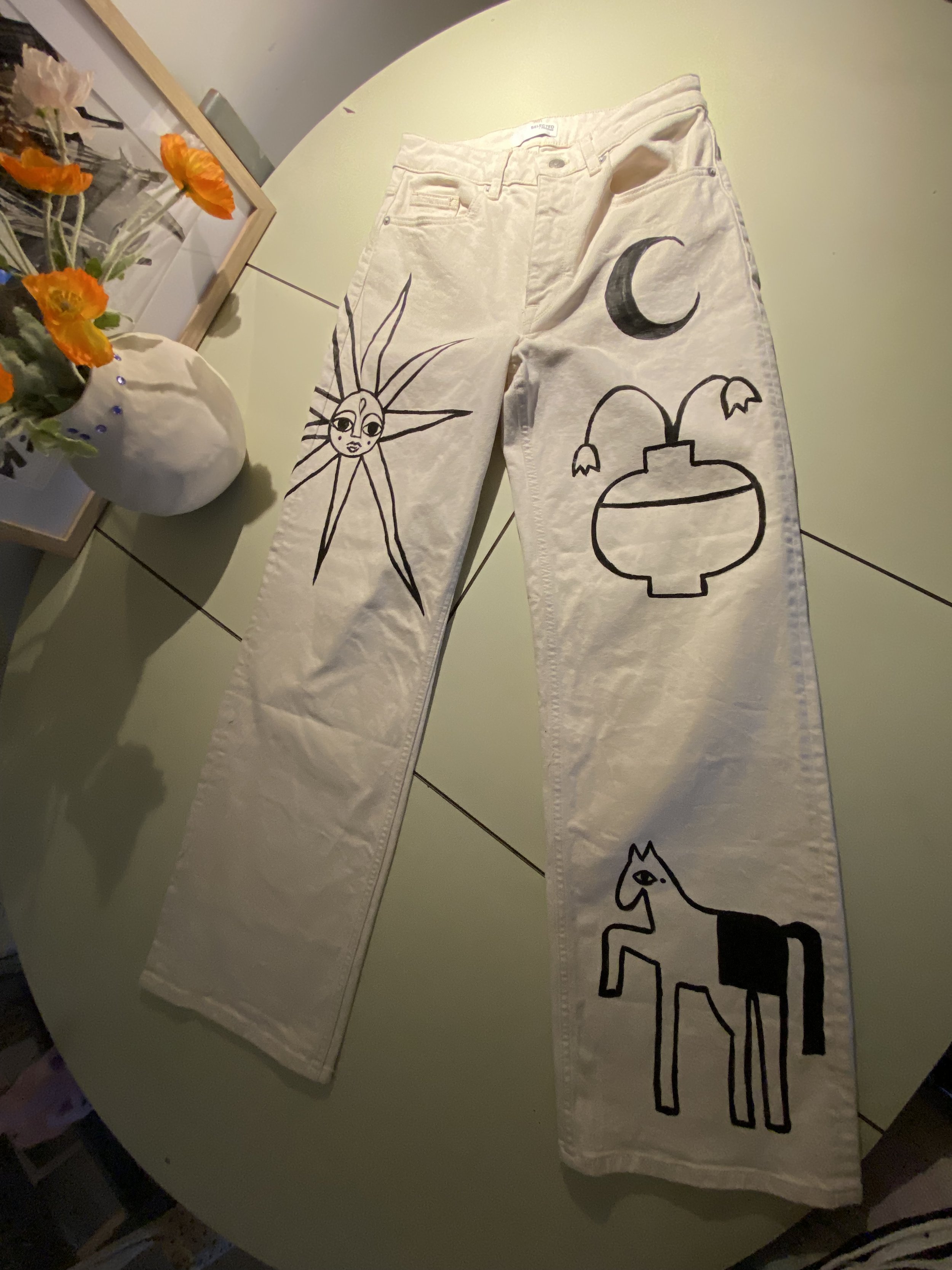 White jeans with black line art designs including a sun with a face, a crescent moon, a lotus flower, and a stylized horse.