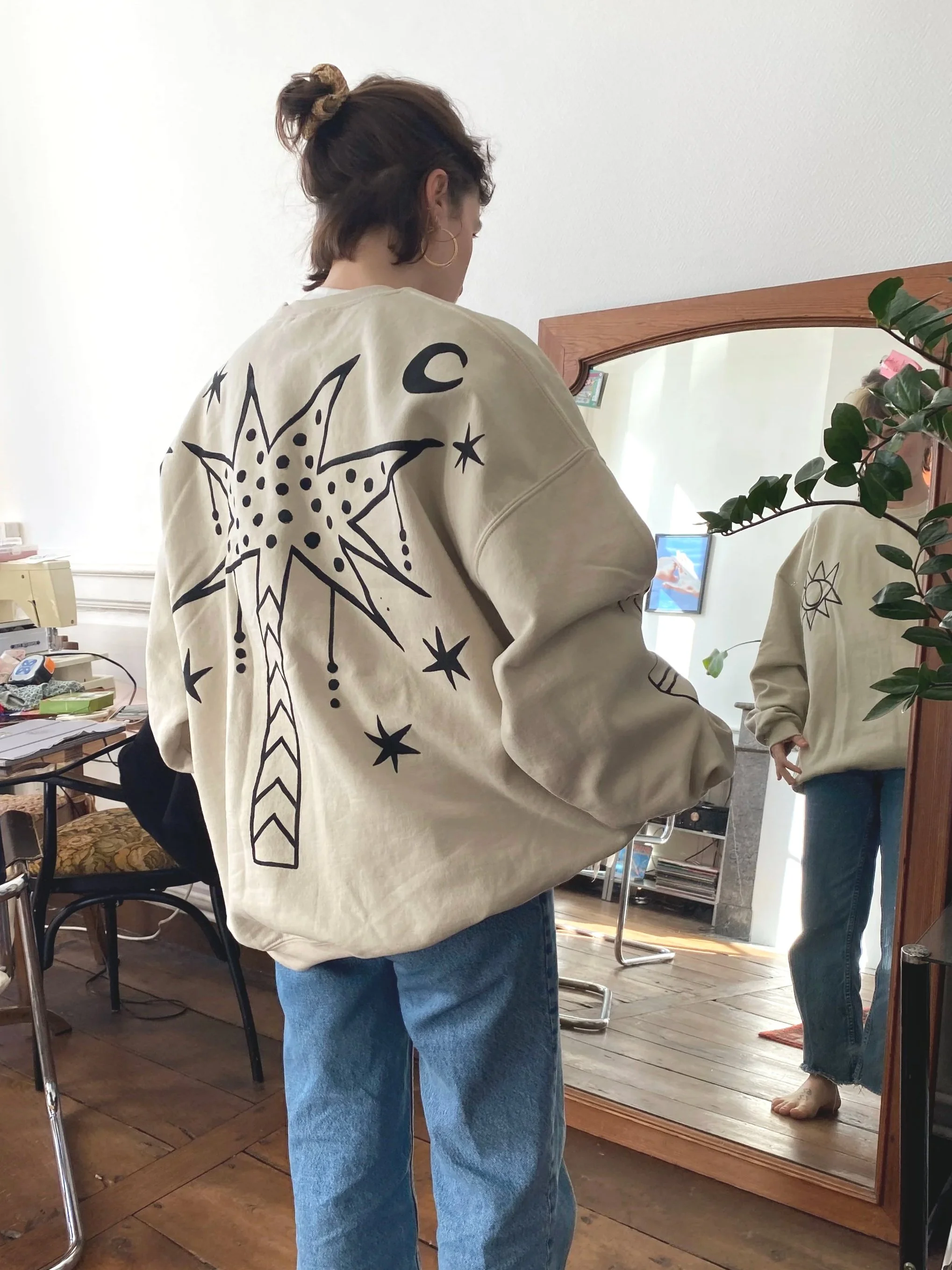 A woman wearing a beige sweatshirt with black celestial and explosion-themed designs, standing in front of a mirror in a room with wooden floors and cluttered desk. She has short hair with a scrunchie, blue jeans, and is looking at her reflection in 