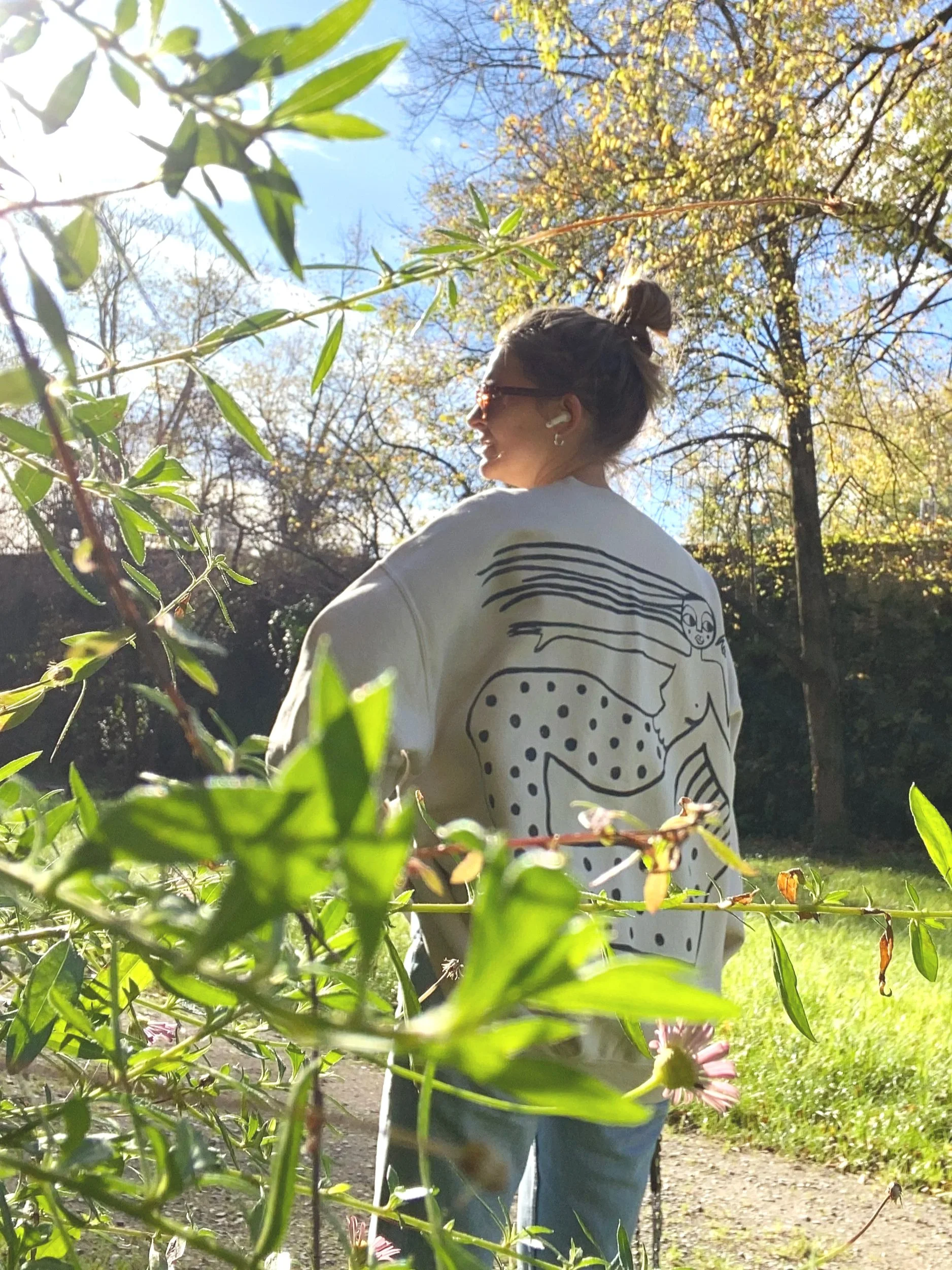 A woman standing outdoors during daytime, wearing sunglasses, earbuds, a white jacket with a whimsical black line drawing of a woman on the back, and light blue jeans, surrounded by trees with some green and yellow leaves, under a clear blue sky.