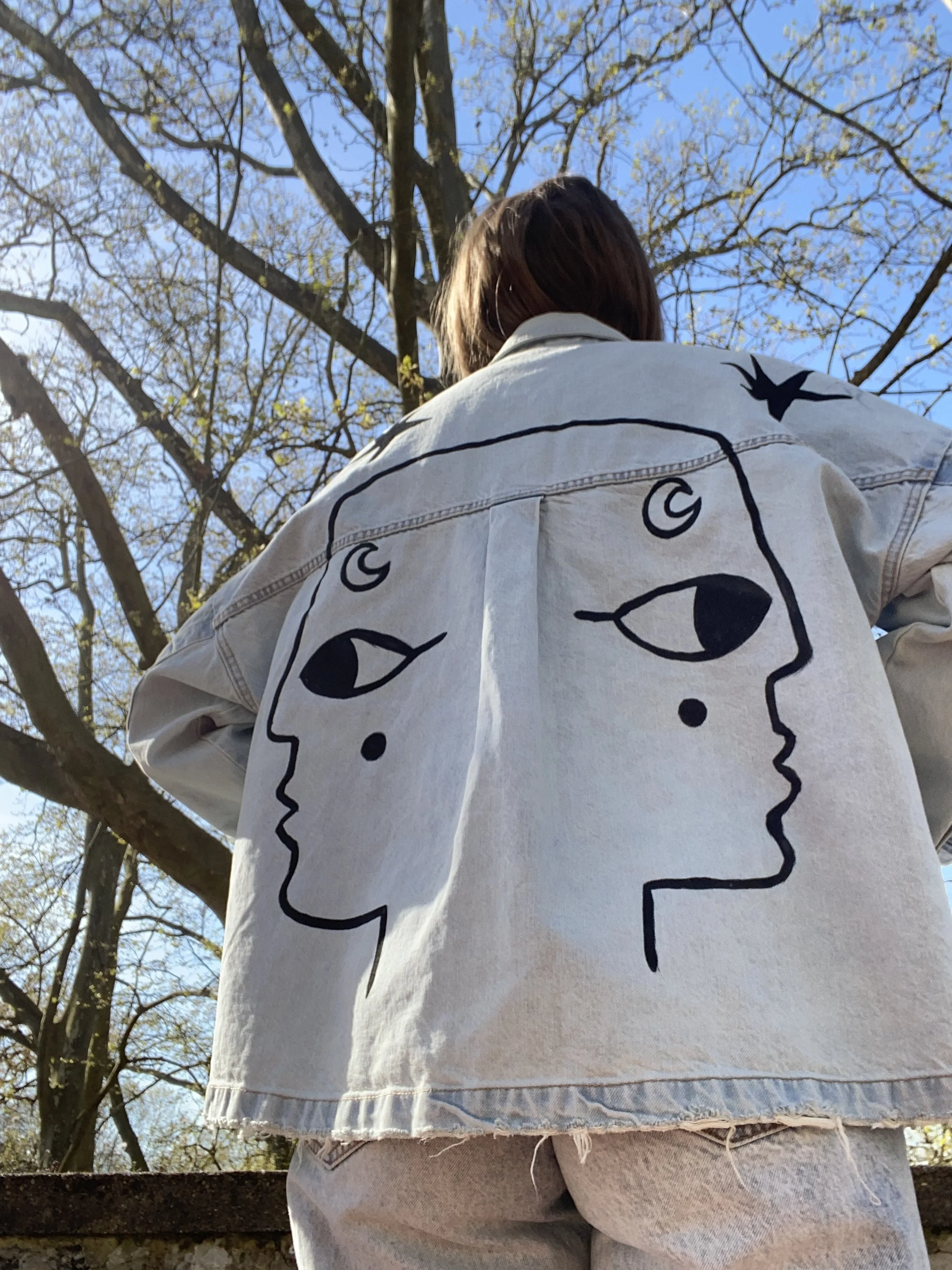 Person wearing a denim jacket with artistic face drawings on the back, standing outdoors under a clear blue sky with tree branches.