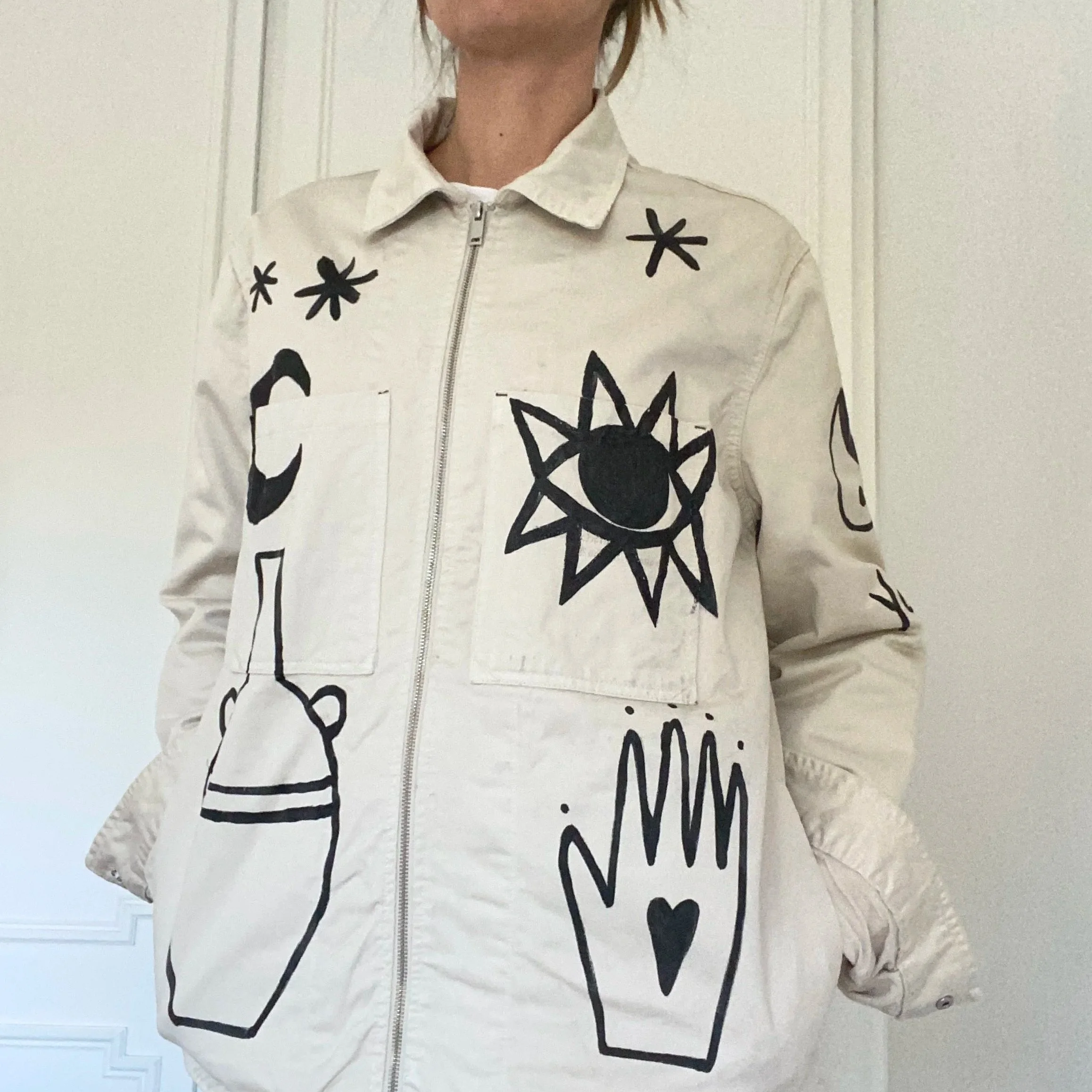 A person wearing a cream-colored jacket decorated with black abstract drawings, including a sun, a vase, a hand with a heart, and stars.