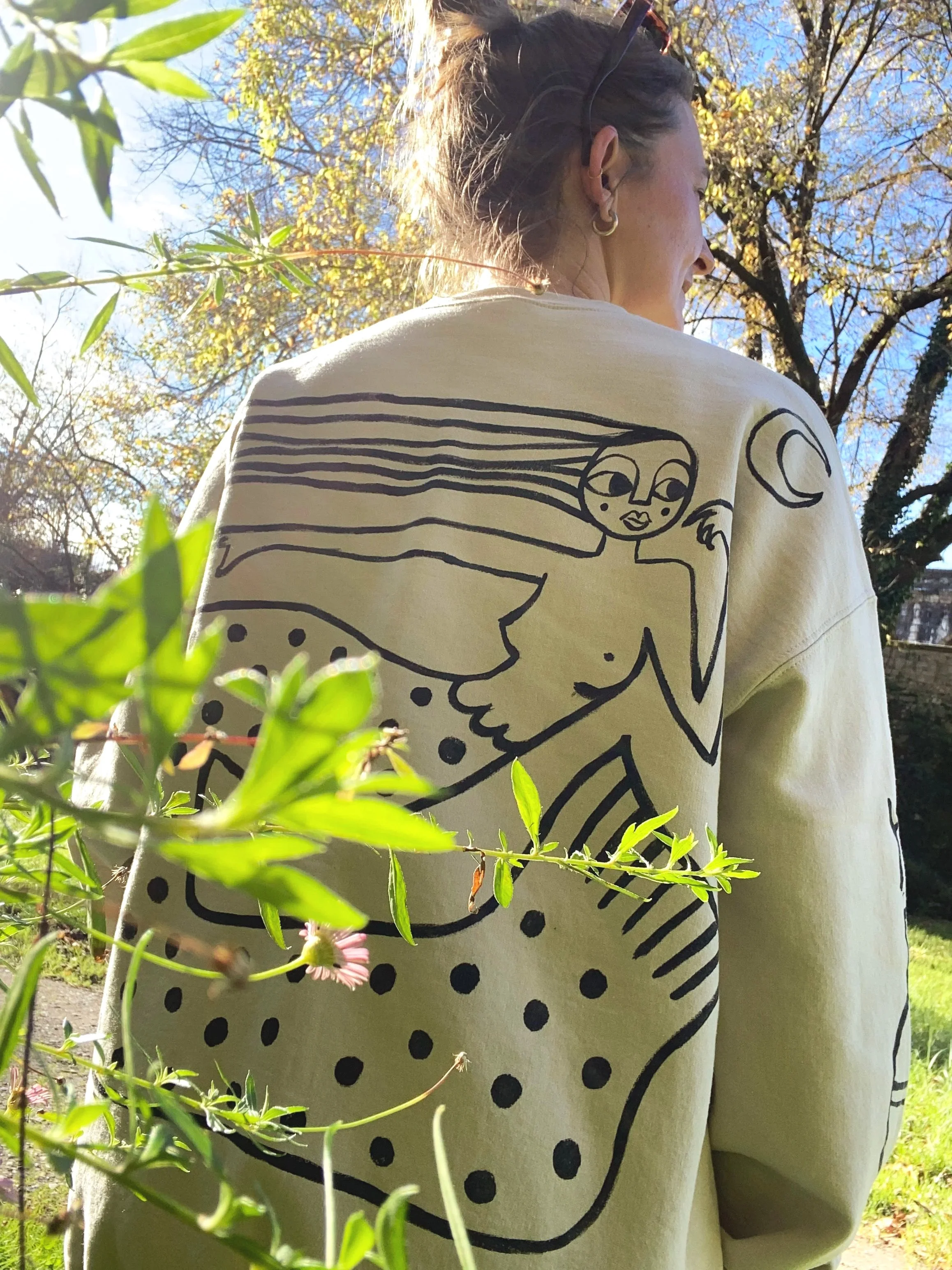 A woman with brown hair tied up, wearing earrings and sunglasses on her head, stands outdoors on a sunny day. She is wearing a cream-colored sweatshirt with a black abstract line drawing of a woman with flowing hair, a moon, and a galaxy on the back.