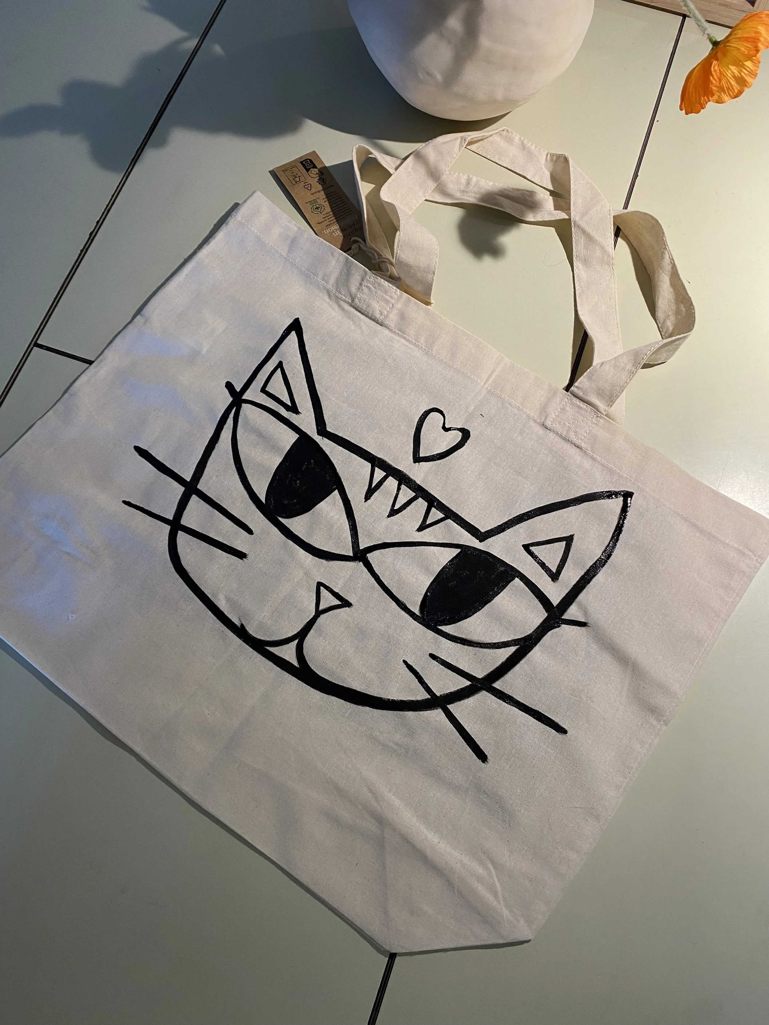 White tote bag with a black line drawing of a cat's face, nose, and whiskers, with a heart above its head, placed on a table with a vase and a flower in the background.