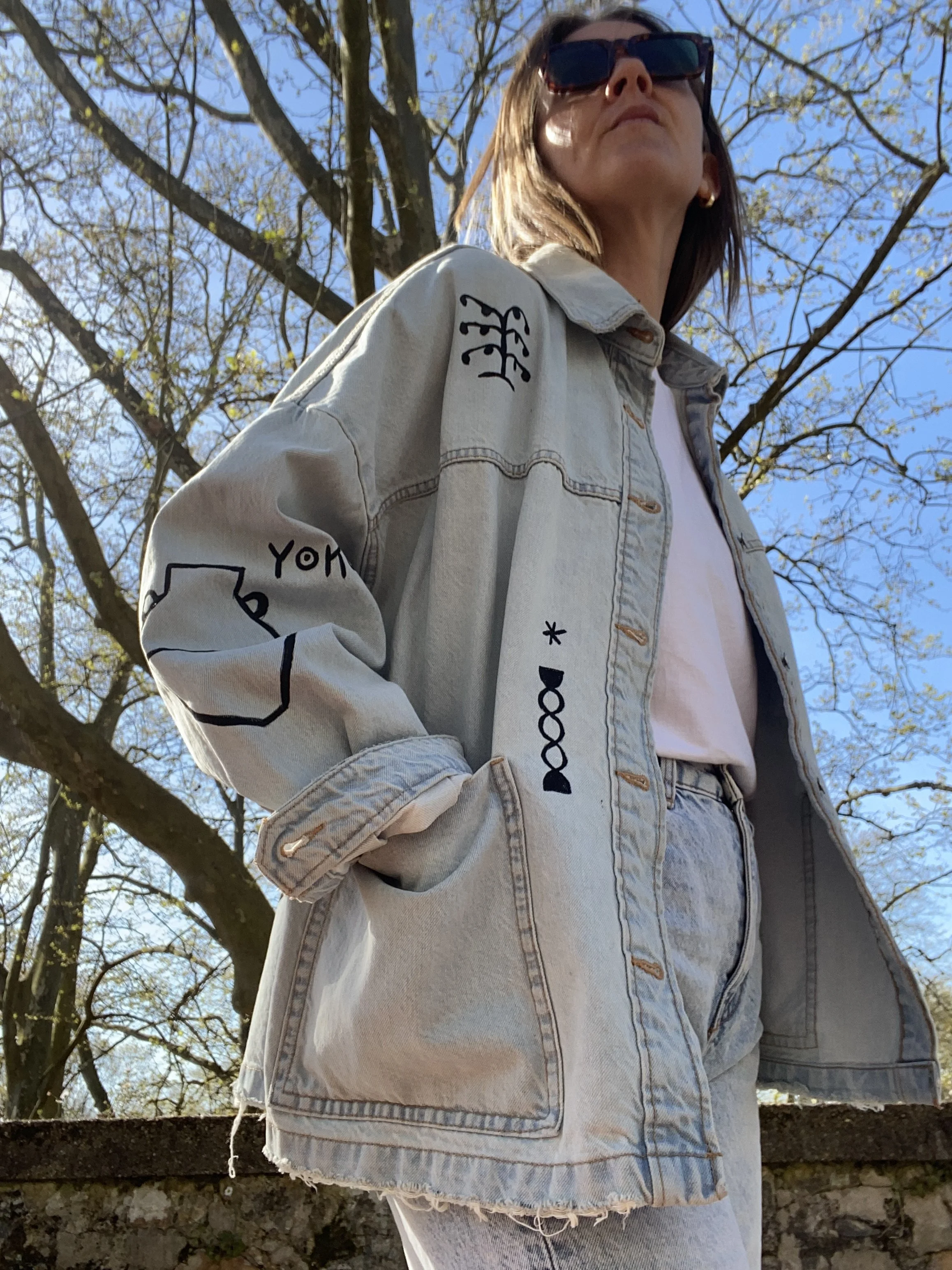 A woman wearing sunglasses, a light-colored denim jacket with black doodle-like designs, and light-colored jeans, standing outdoors under a tree with budding leaves and a clear blue sky.