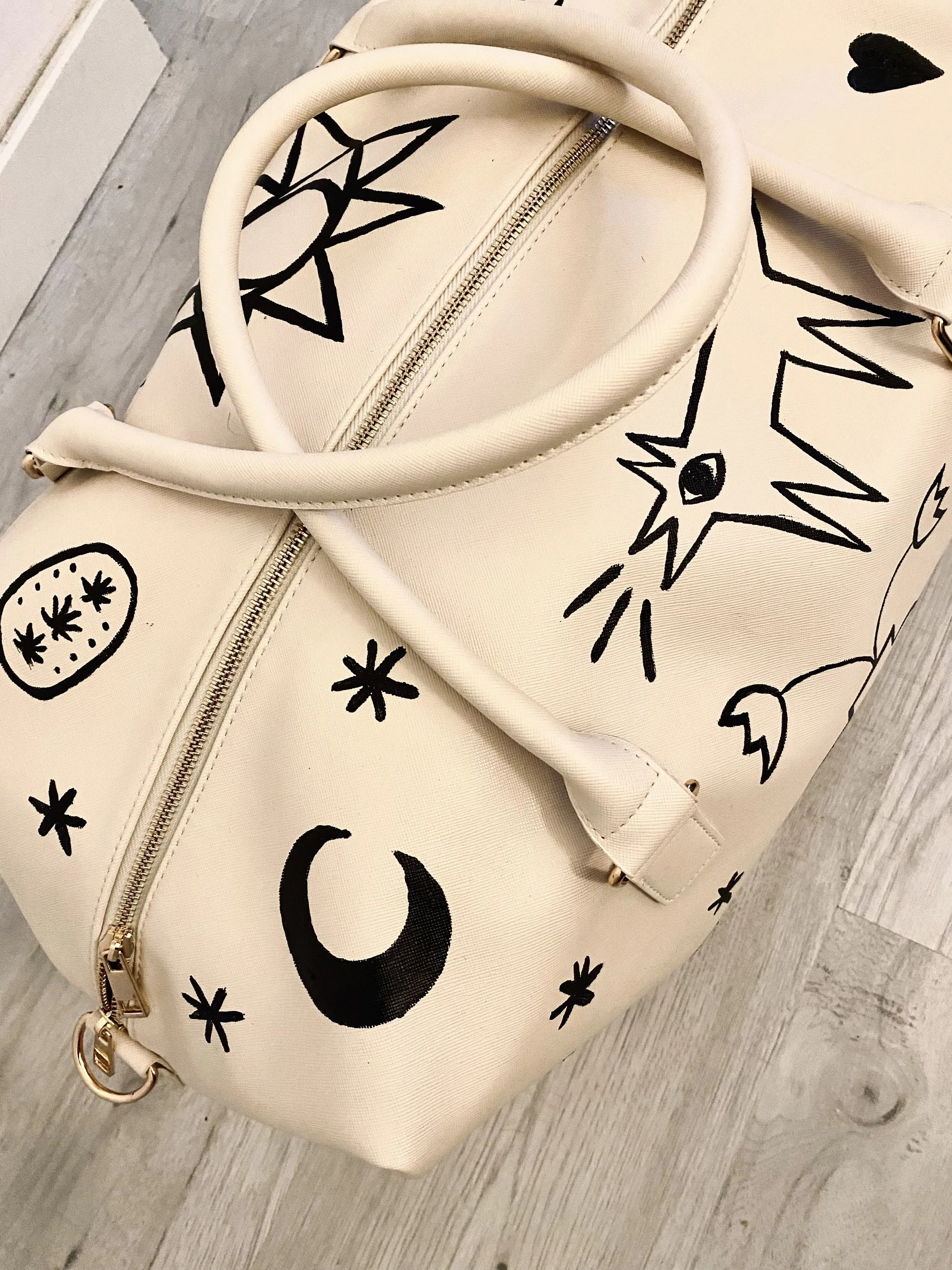 Cream-colored handbag with black doodle-style drawings of celestial and abstract shapes, including stars, moon, and plants, on a wooden floor.