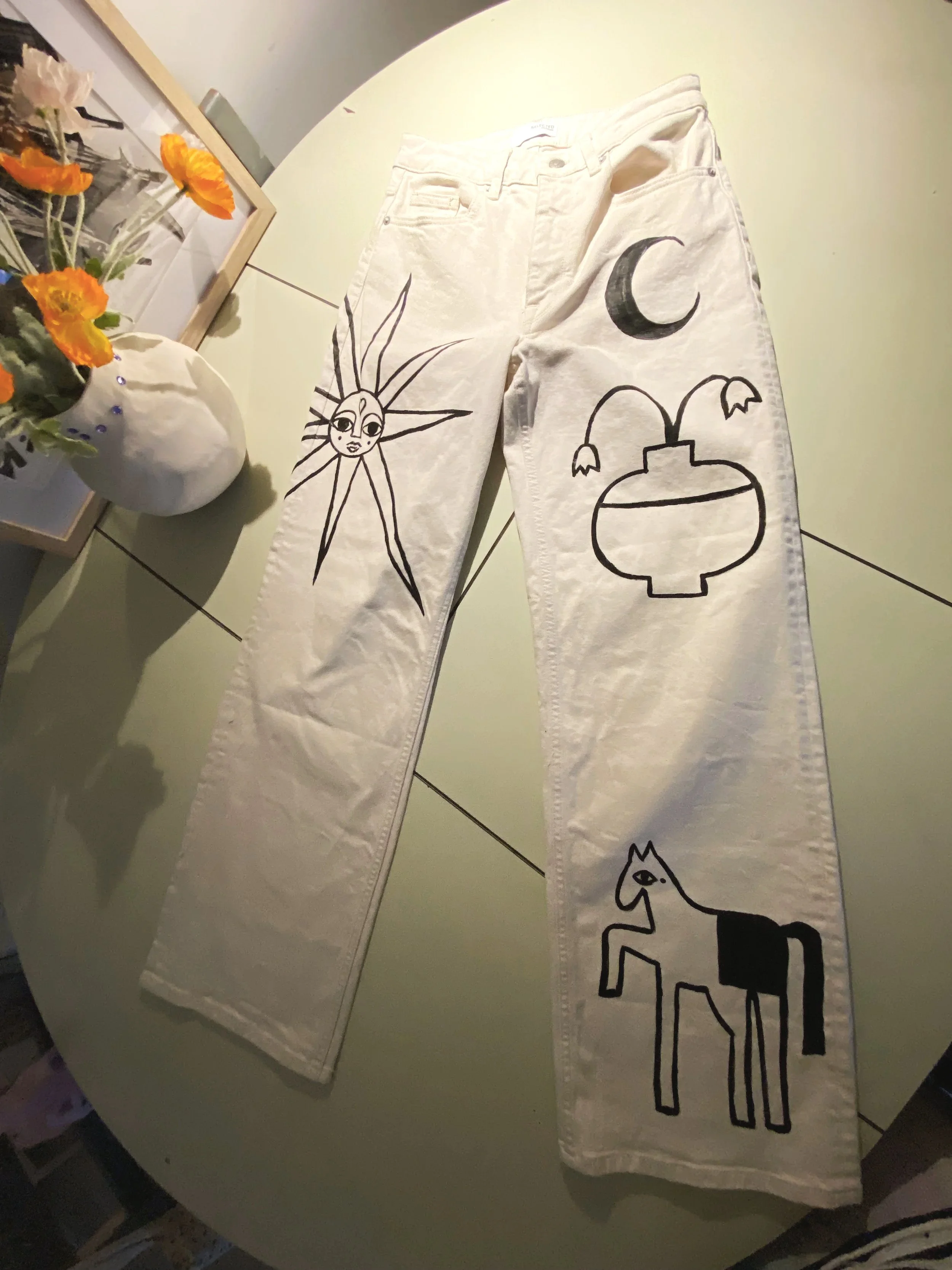 White jeans with black line drawings of a sun with a face, a crescent moon, a vase with flowers, and a stylized horse, laid out on a table with a vase of orange flowers nearby.