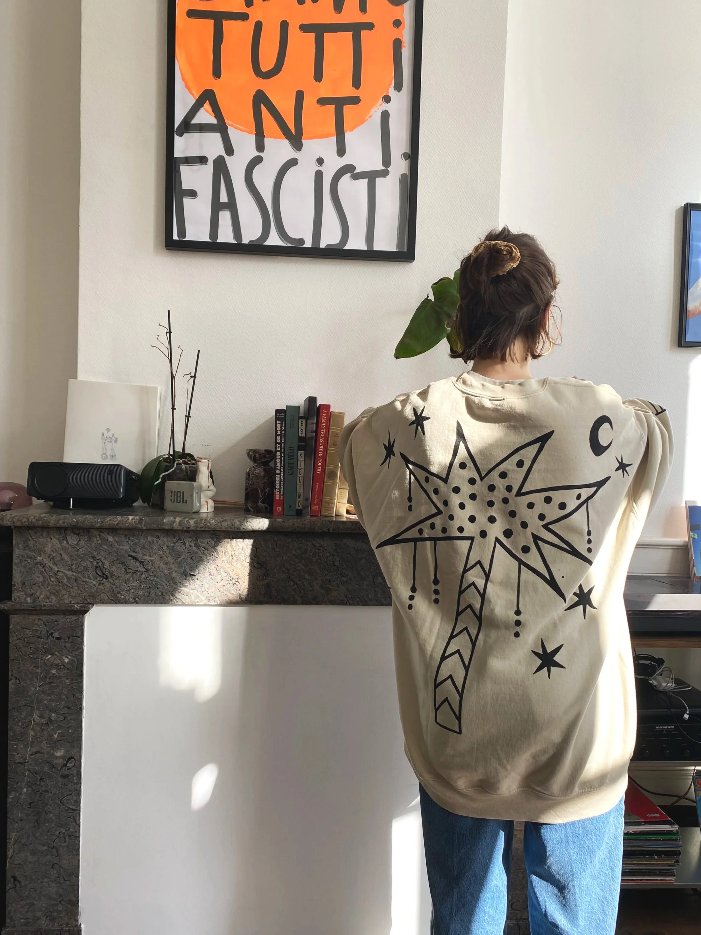 A person with short hair in a beige sweatshirt with a black outlined star and moon design on the back, standing with their back to the camera in a room with sunlight. They are facing a countertop with books, a plant, and a JBL speaker. A framed poste