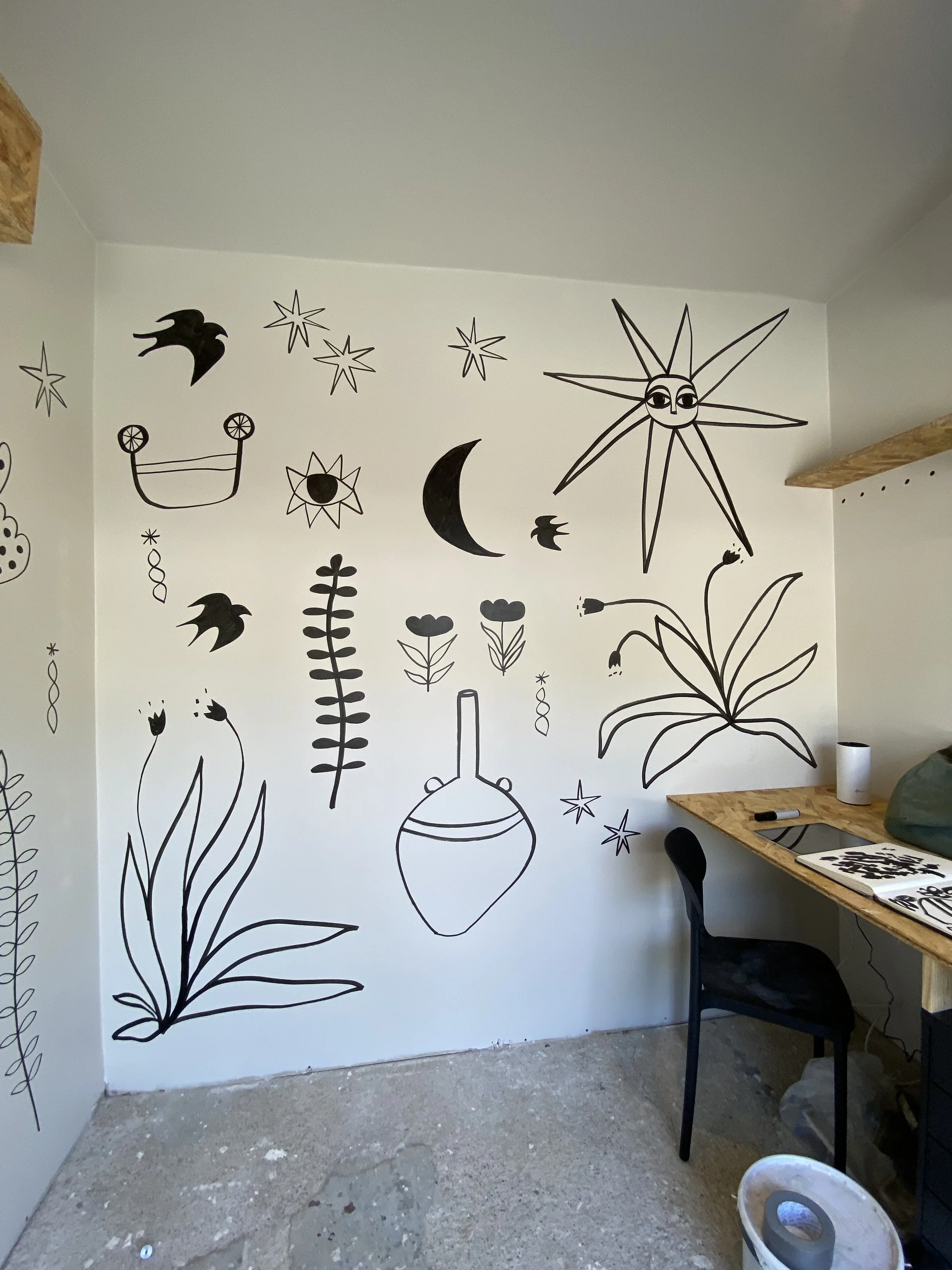Black line art of celestial and botanical elements including stars, a crescent moon, a sun with a face, birds, flowers, and a vase on a white wall.