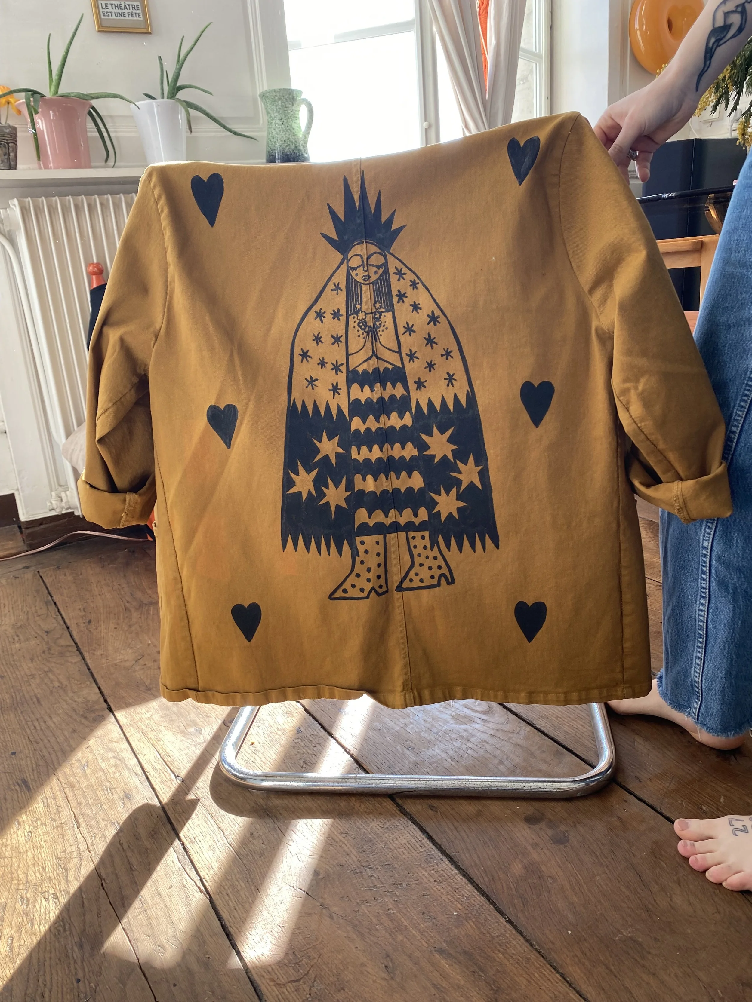 A mustard yellow jacket with an artistic illustration of a woman with a crown, hands pressed together, surrounded by four black hearts, hanging on a chair. The setting appears to be a cozy indoor space with wooden floors, potted plants, and sunlight coming through a window.