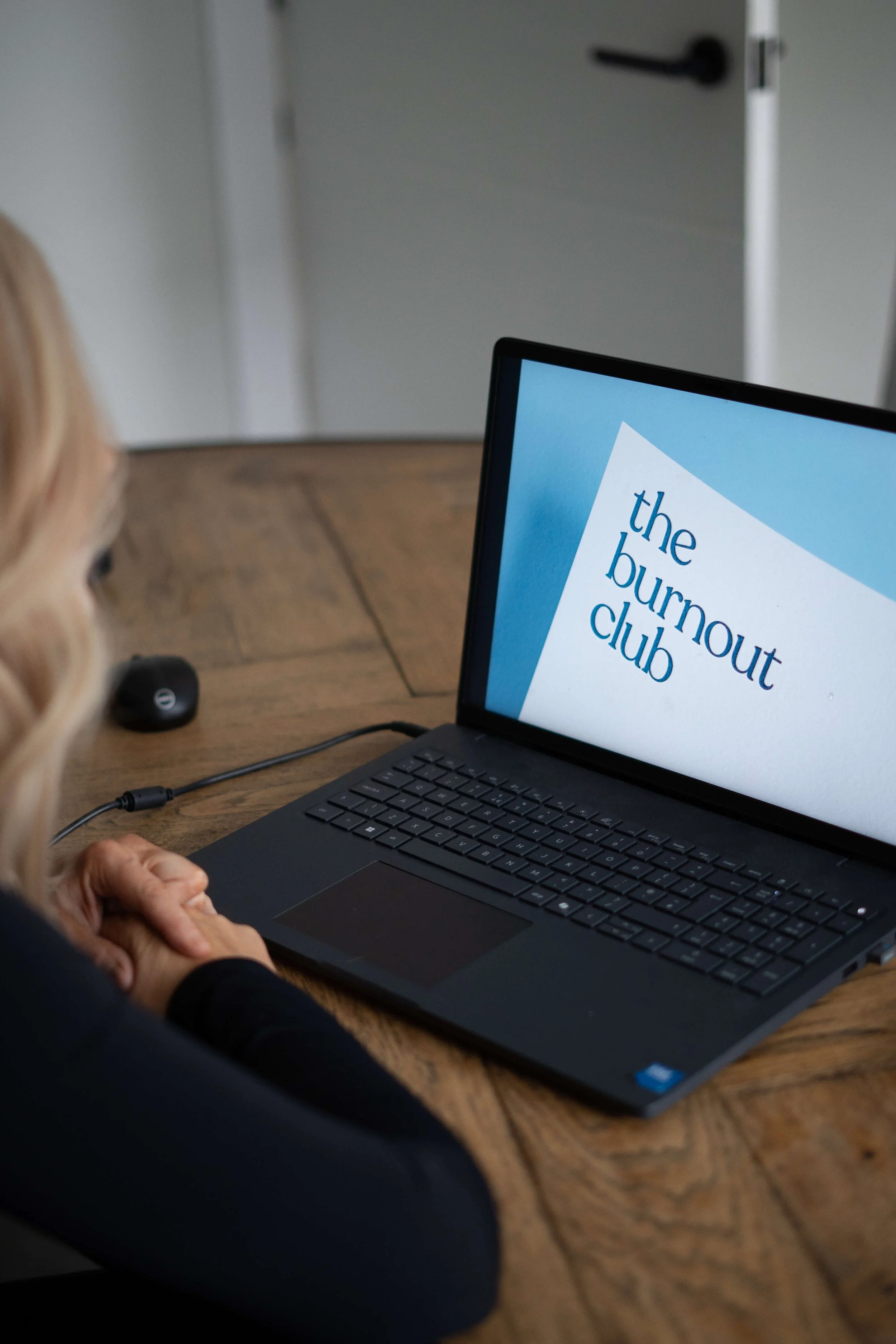 A woman with blonde hair sits at a wooden table looking at a laptop screen that displays the logo 'the burnout club.'