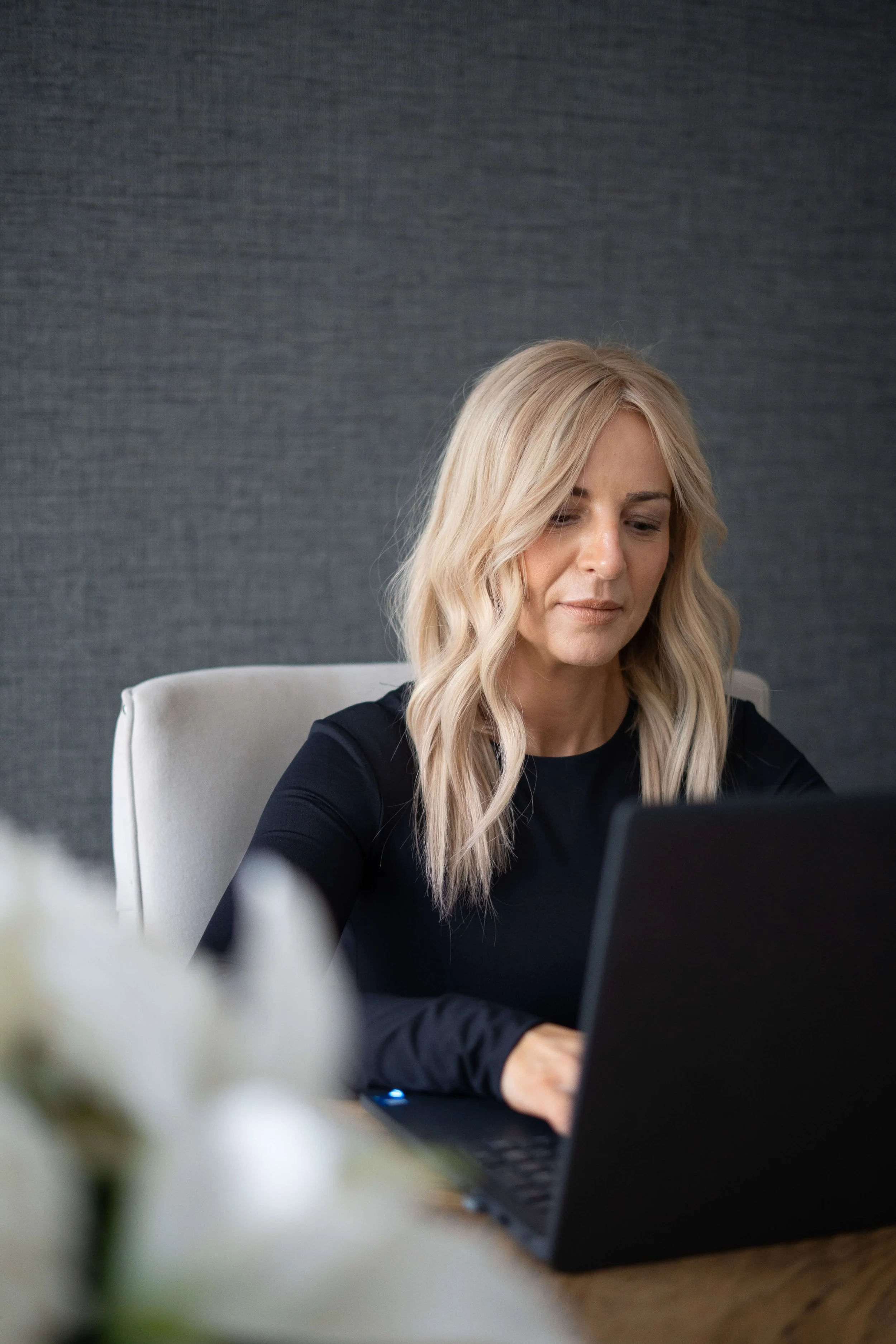 A woman with blonde hair in loose waves, wearing a black top, working on a laptop at a desk with a dark textured wall in the background.
