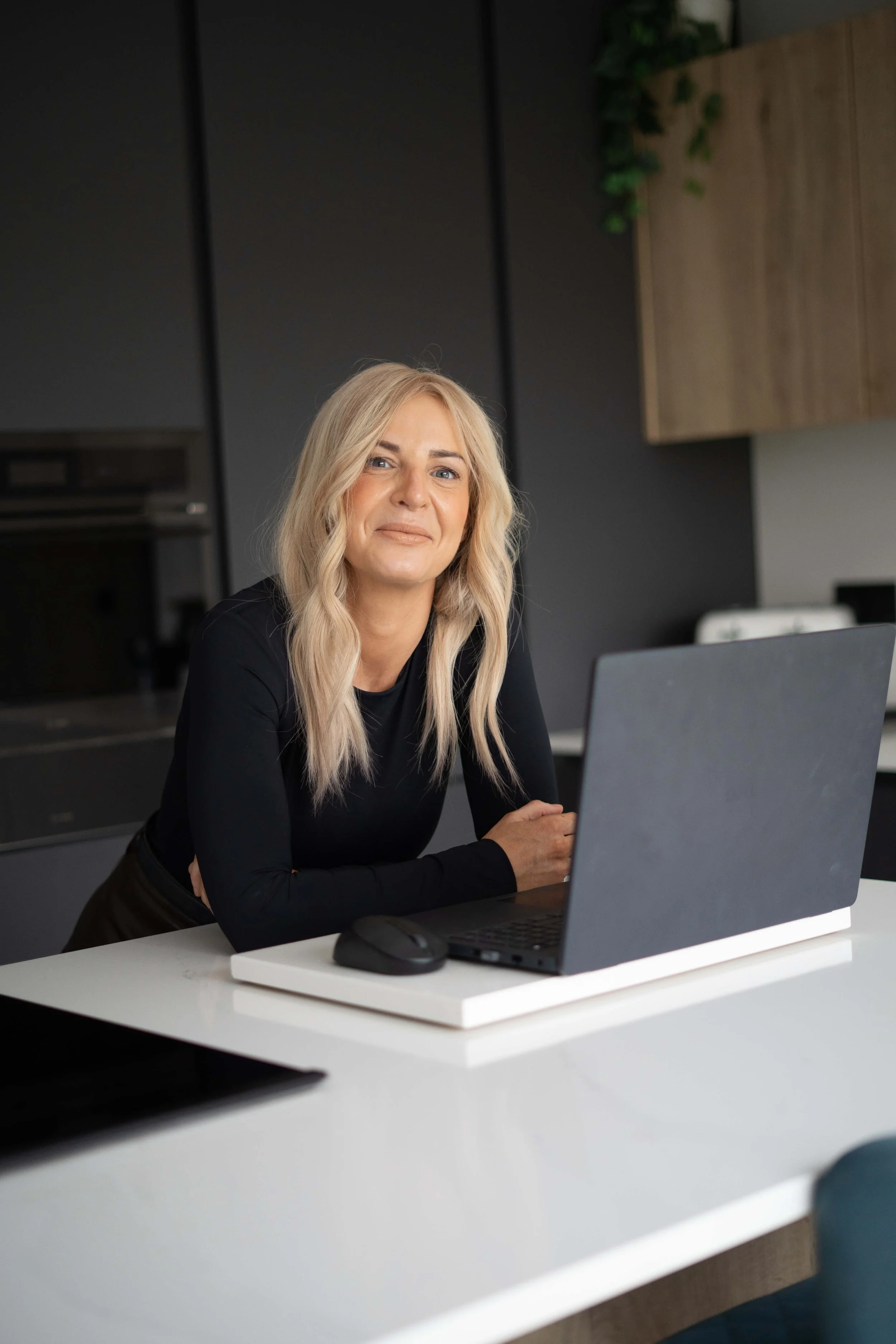 A woman with blonde hair and a black top sitting at a white desk with a laptop and mouse, smiling at the camera in a modern kitchen or office setting.