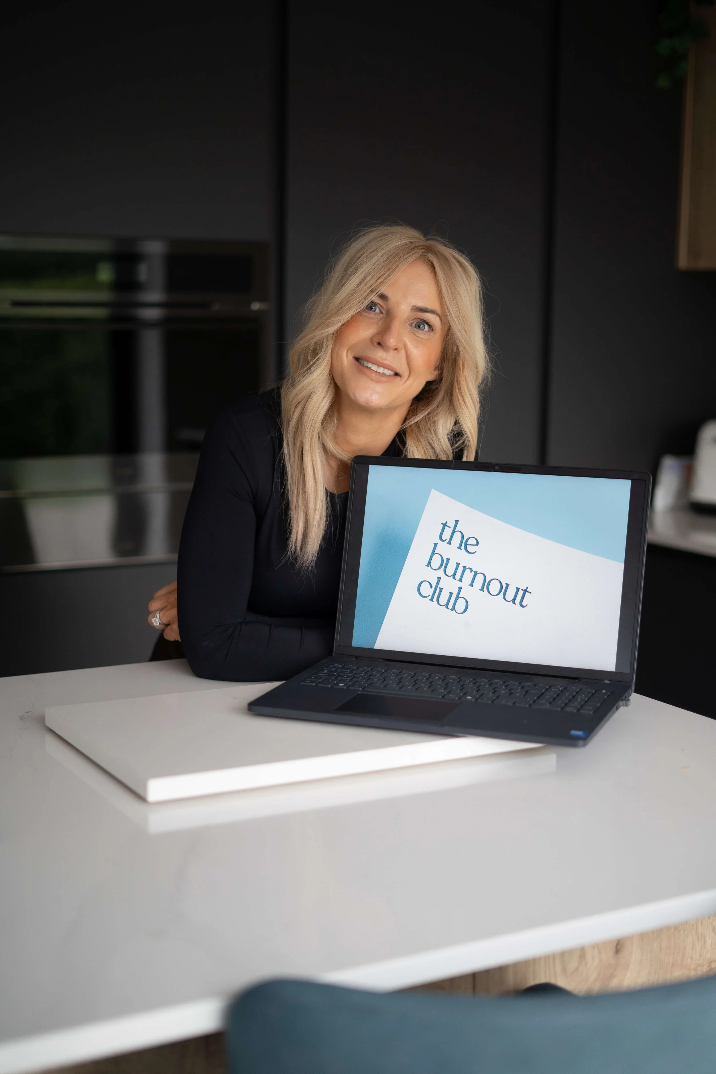 A woman with blonde hair smiling while sitting at a kitchen table next to a laptop displaying a slide with the title 'the burnout club.'