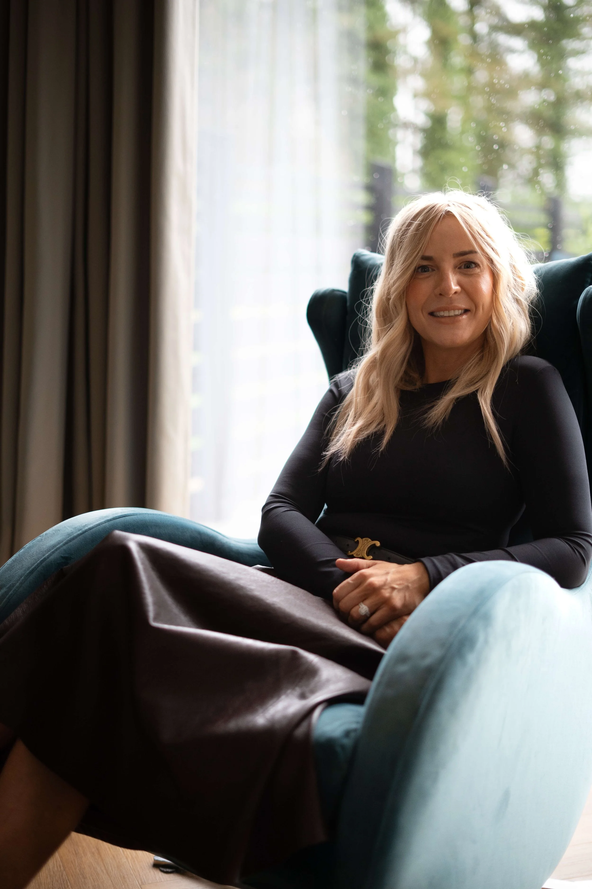 A woman with blonde hair sitting in a teal armchair in front of a window with curtains, smiling at the camera.