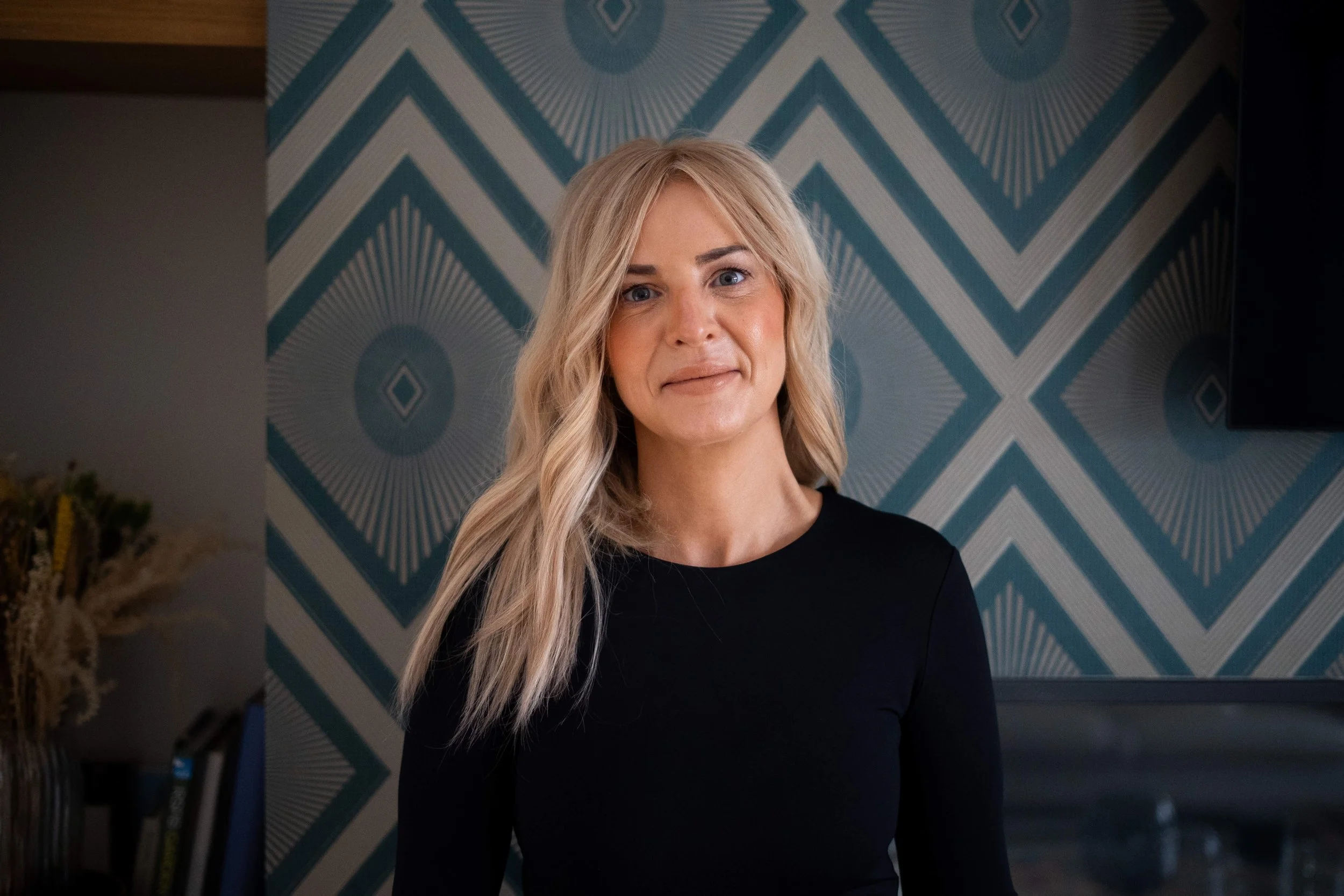A blonde woman with shoulder-length wavy hair wearing a black top, standing in front of a geometric patterned blue and gray wall.