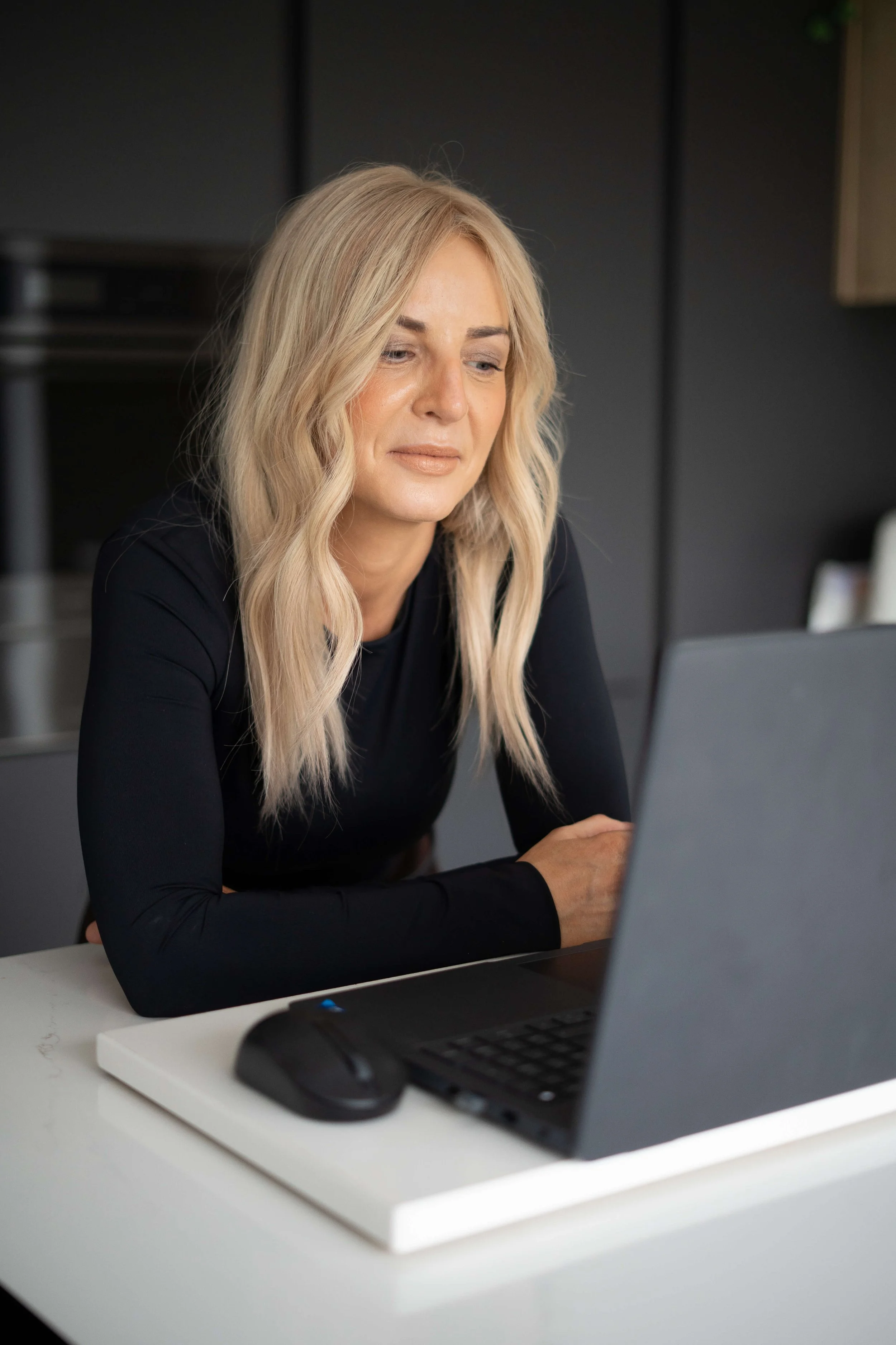 Woman with blonde hair sitting at a desk, looking at a laptop.