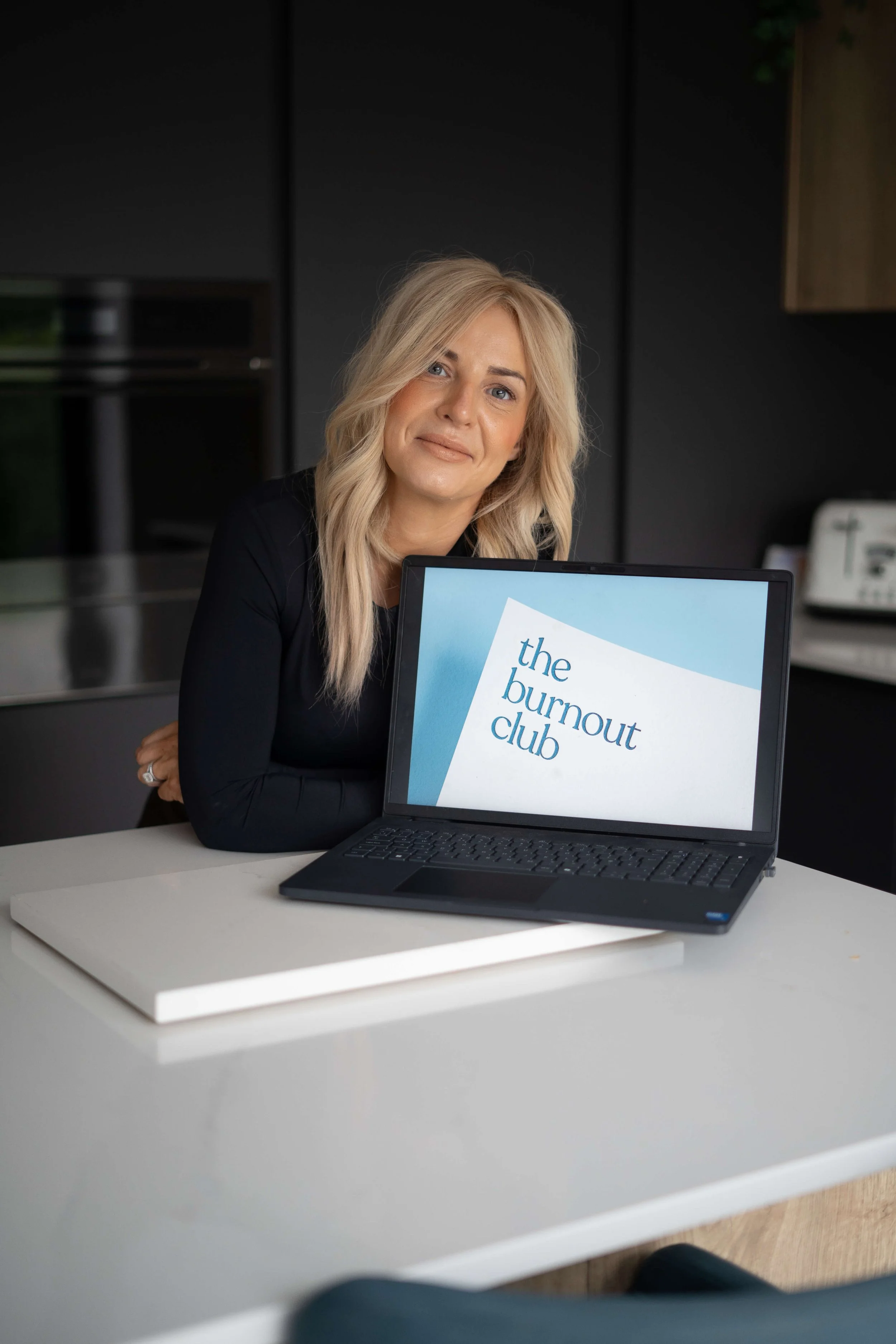 A woman with blonde hair sitting at a white table with a laptop displaying the text "the burnout club"