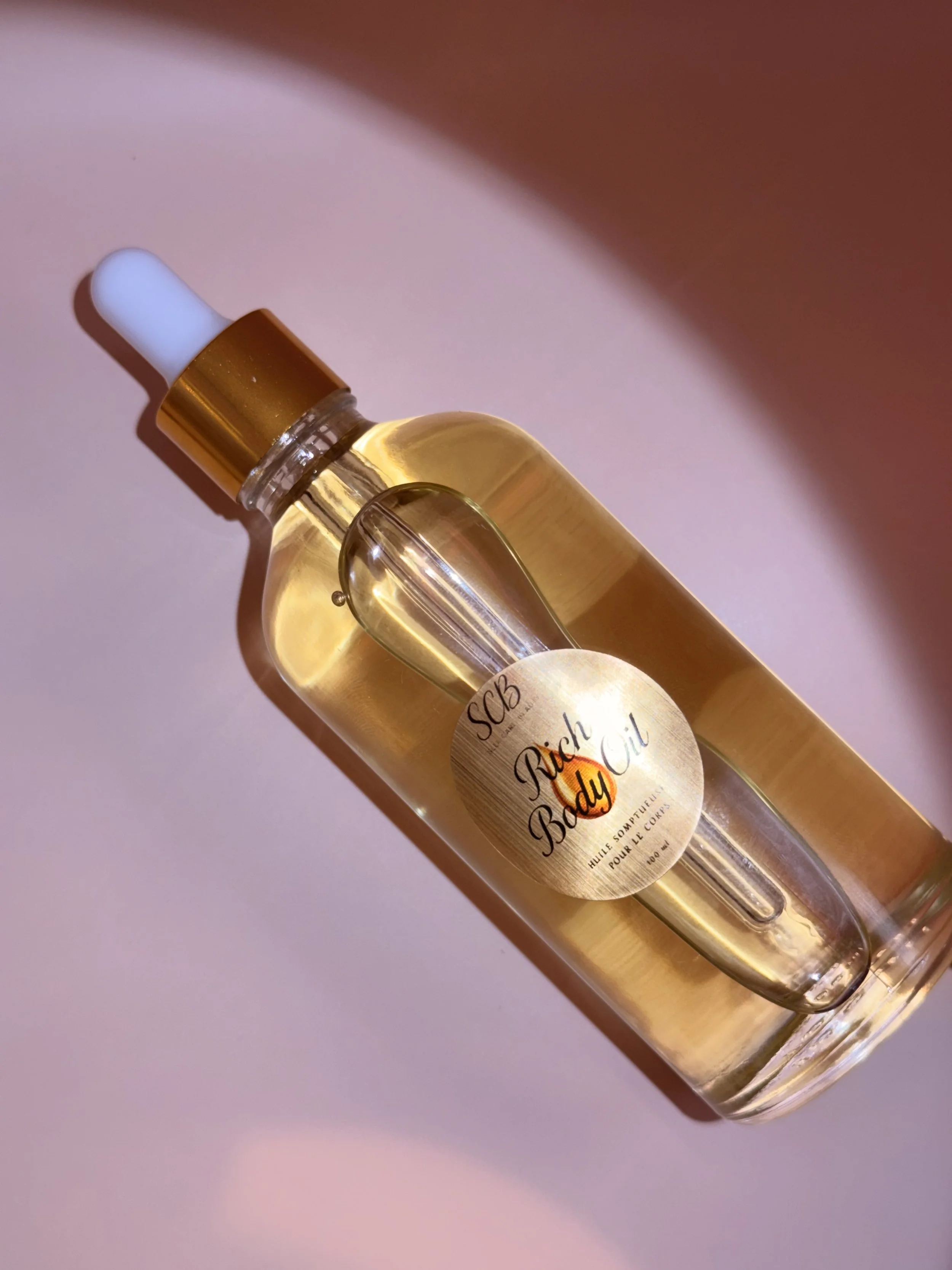 Une bouteille d'huile pour le corps appelée 'Rich Body Oil', avec un bouchon doré et un flacon en verre clair, placé sur une surface rose pâle avec un éclairage créant une ombre douce.