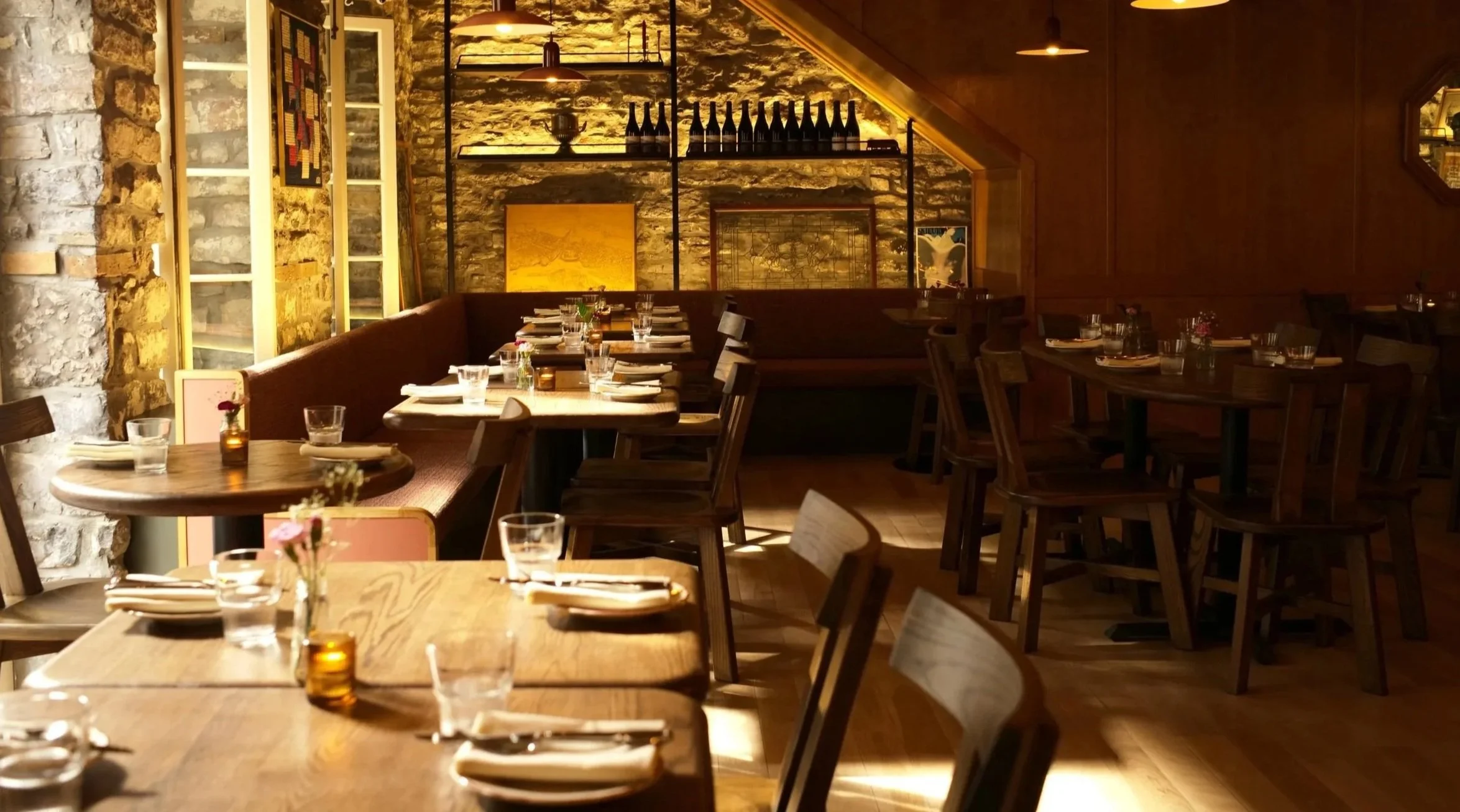 Interior of a cozy restaurant with wooden tables and chairs, stone walls, and warm lighting, set for dining with glasses, napkins, and small vases with flowers.