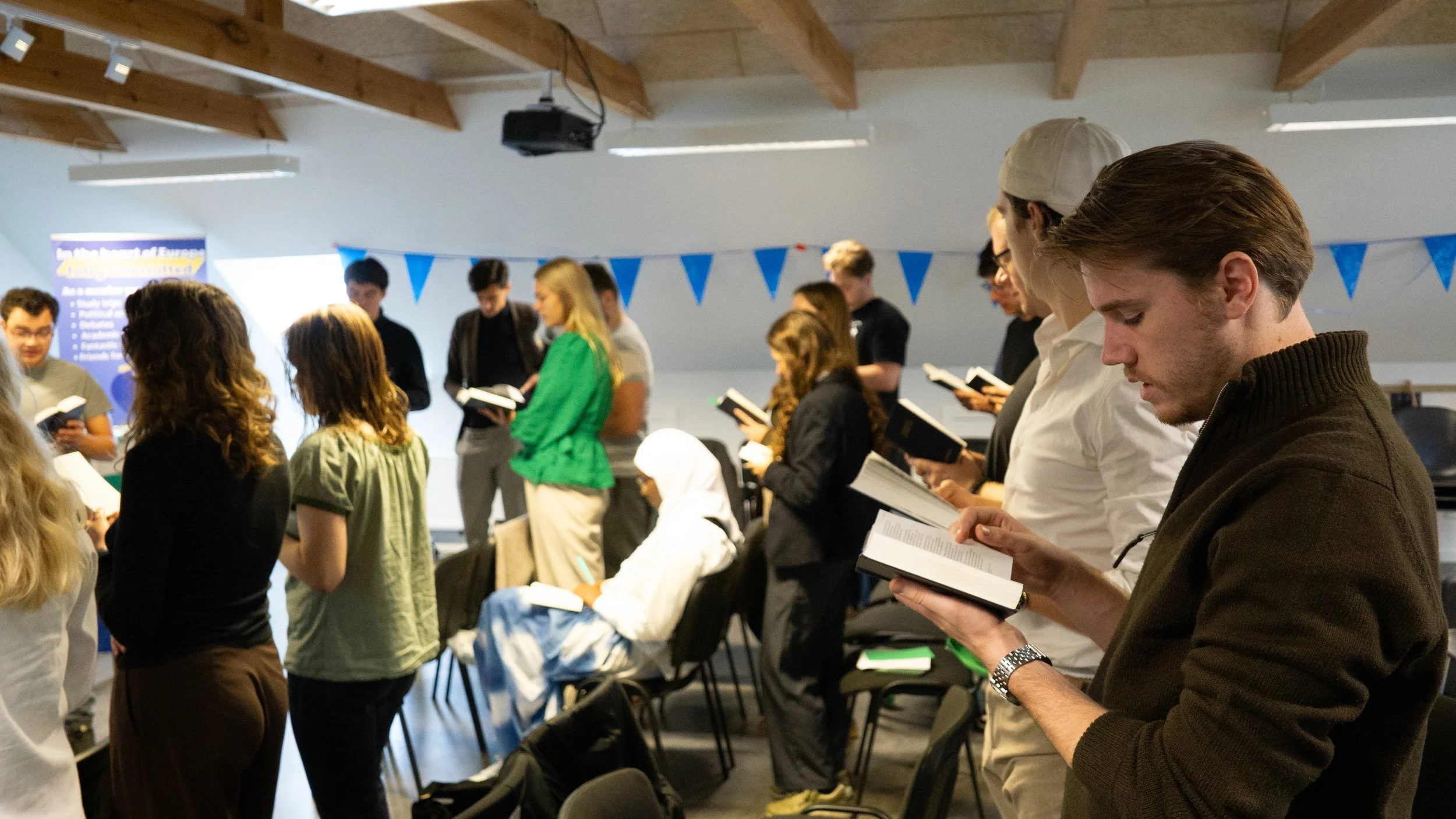 En gruppe unge mennesker står og læser i bøger i et røgfrit indendørs rum med træbjælker i loftet og blå flagdekorationer.