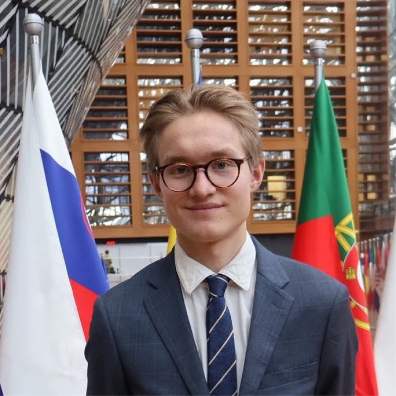 En ung mand i jakkesæt og slips, med brunt hår og briller, står foran flere europæiske flags.