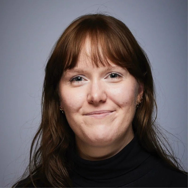 Portræt af en kvinde med brunt hår, iført sort turtleneck, smilende mod kameraet, mod en grå baggrund.