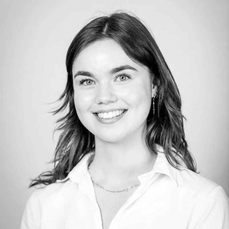 Sort-hvid portrætfoto af en smilende ung kvinde med skulderlangt hår, iført en hvid skjorte og smykker.