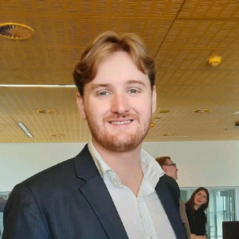 En ung mand med let skæg og lyst hår iført en hvid skjorte og mørk jakkesæt, smilende ved en indendørs begivenhed.