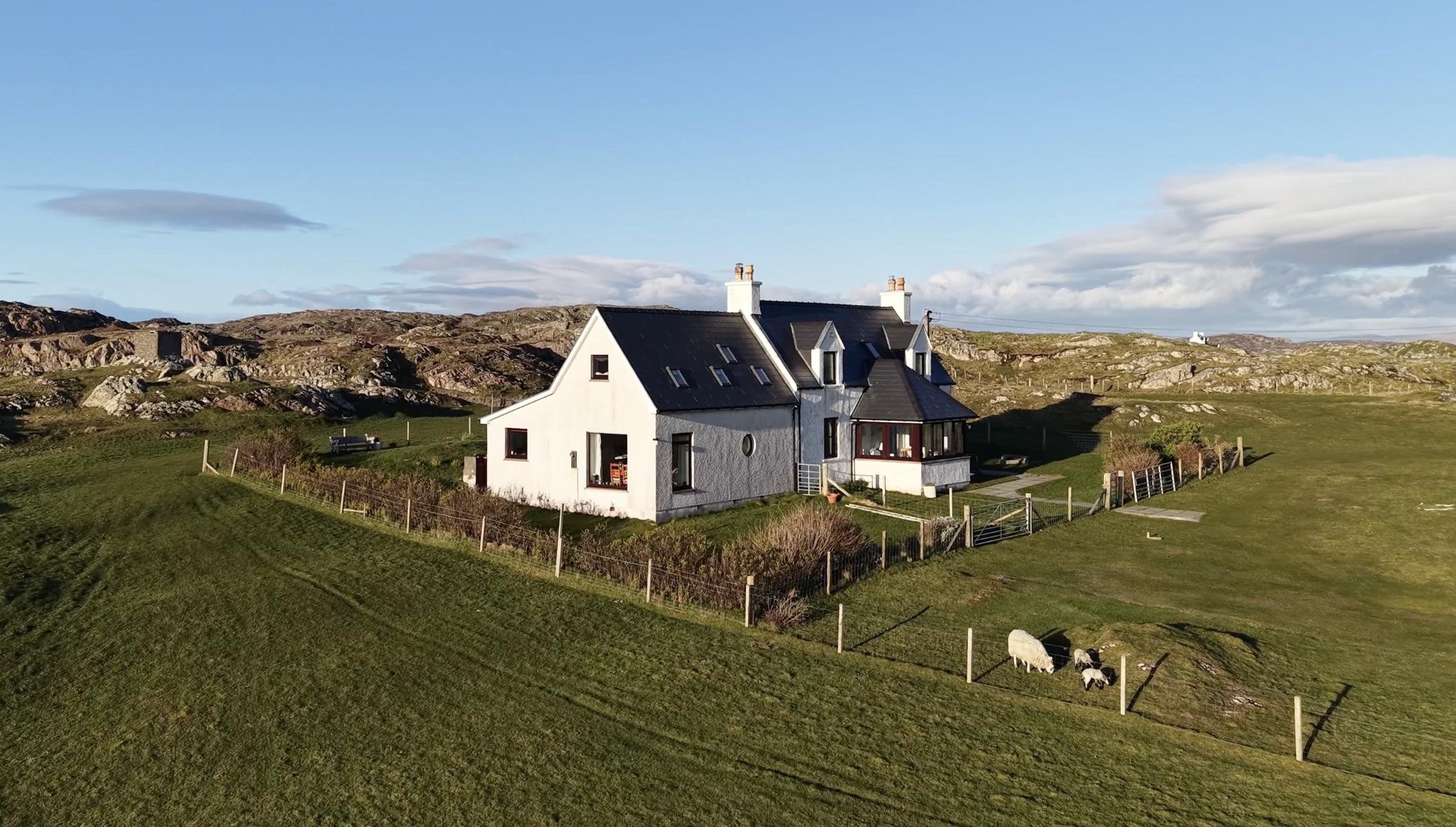 A white house - known as Machair House - with black roof and multiple chimneys in a green landscape with hills and sheep grazing, under a partly cloudy sky.