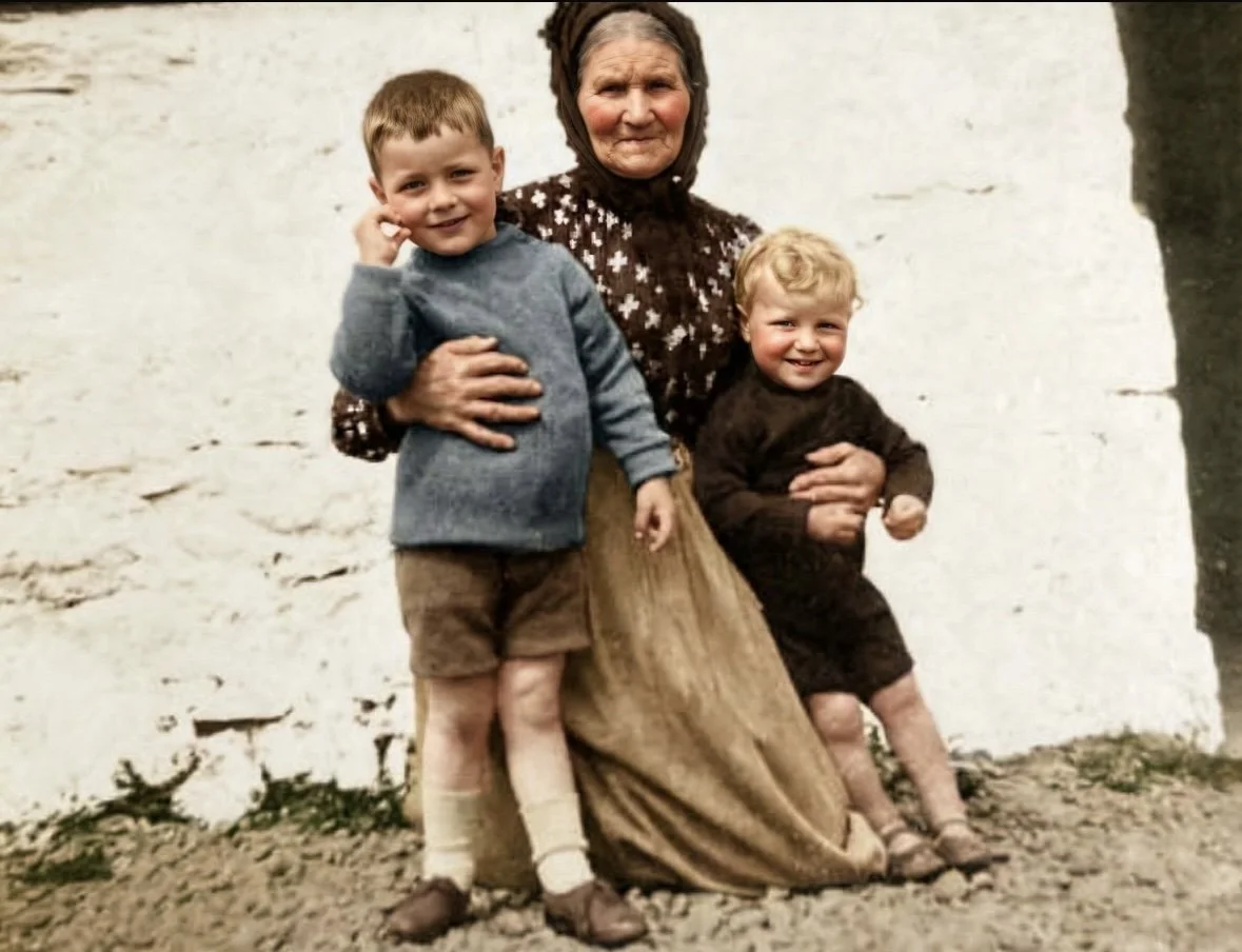 An elderly woman with two young boys outdoors near a white wall, smiling and holding the boys, who are dressed in vintage clothing.