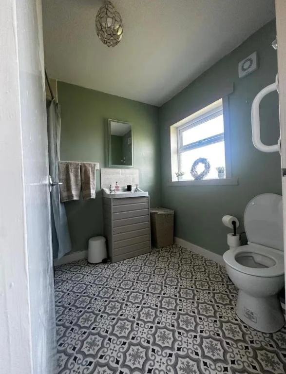 A small bathroom with green walls, a window with a wreath and two small potted plants, a toilet with a cover up, a grey cabinet, a mirror above the sink, a small stool, and patterned floor tiles.