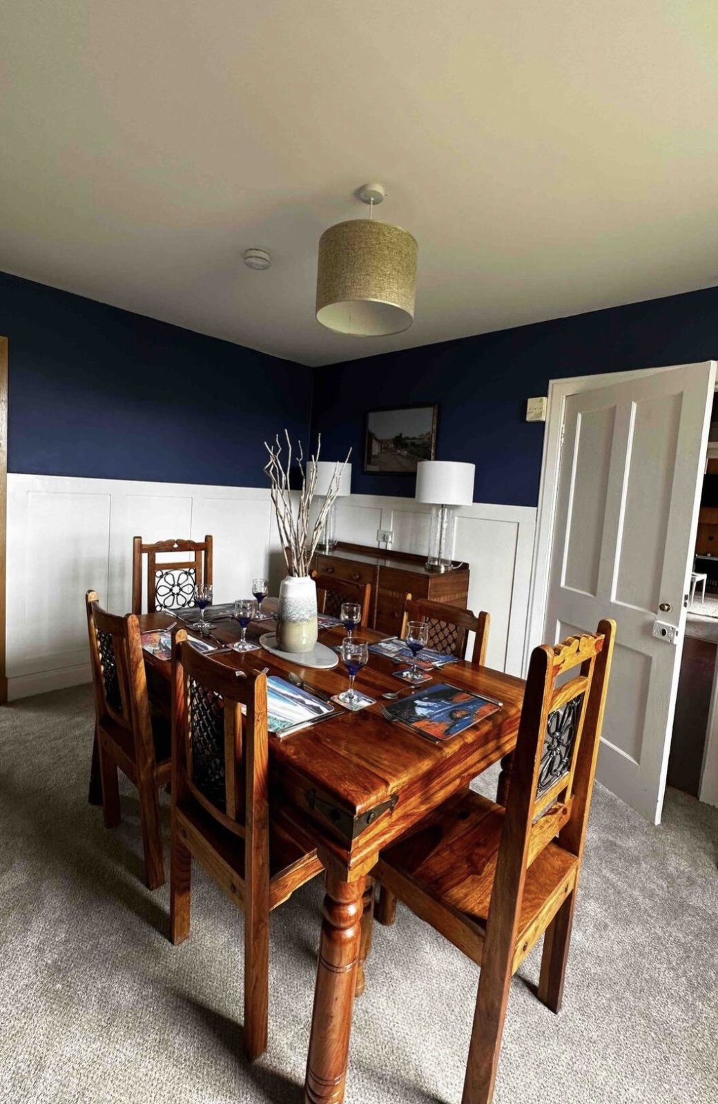 A dining room with a wooden table set for six, decorated with a large vase of white branches as a centerpiece. There are six wine glasses, plates, and utensils arranged around the table. The walls are painted dark blue with white wainscoting, and the