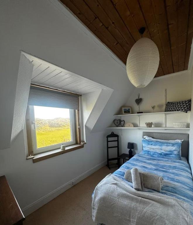 A cozy bedroom with a sloped attic ceiling, a window with a view of green fields, a single bed with blue striped bedding, a small black bedside lamp, a wooden chair, and white shelves with decorative items.